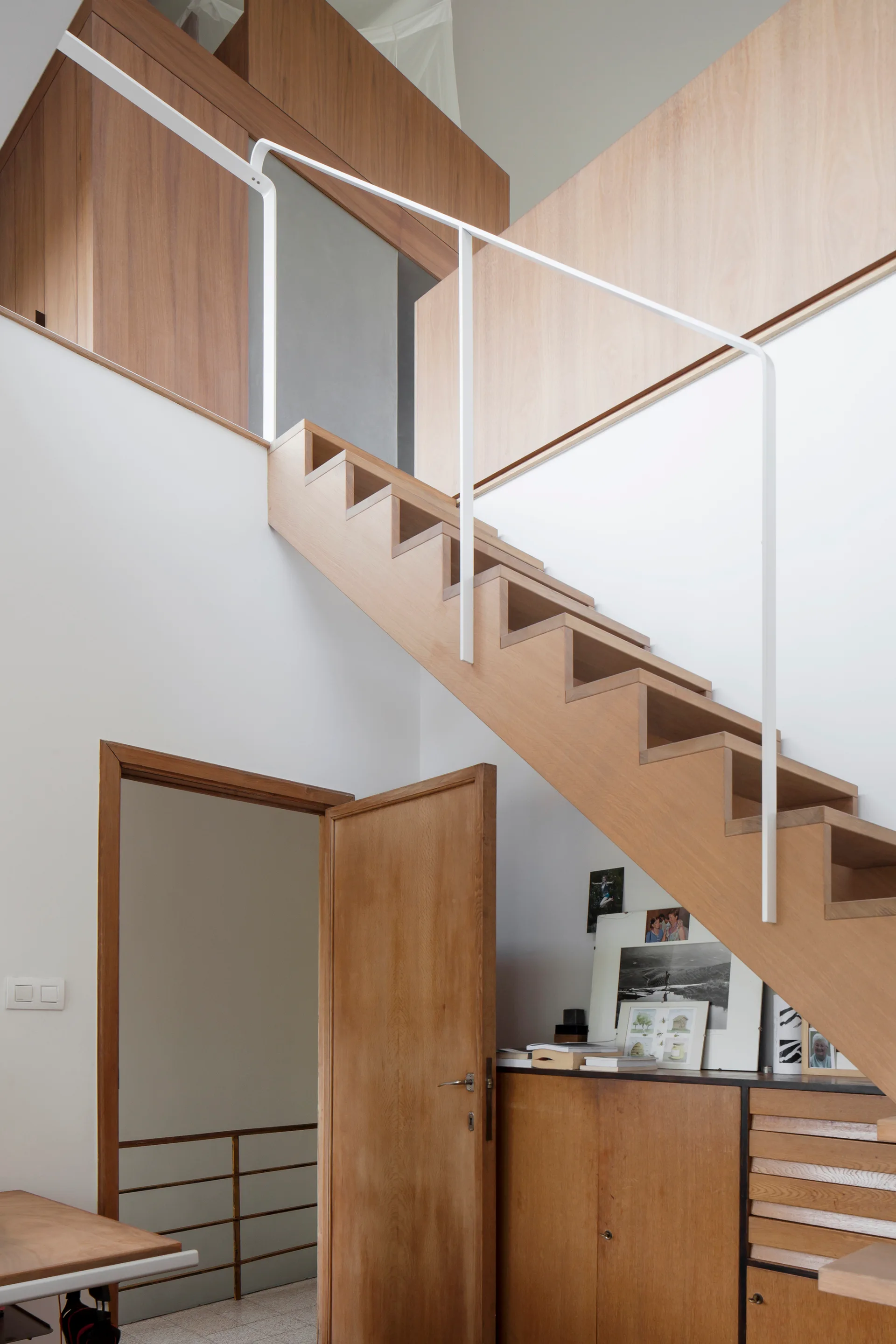 A wooden sawtooth staircase with a slim white handrail stands next to an open wooden door and built-in cabinets.