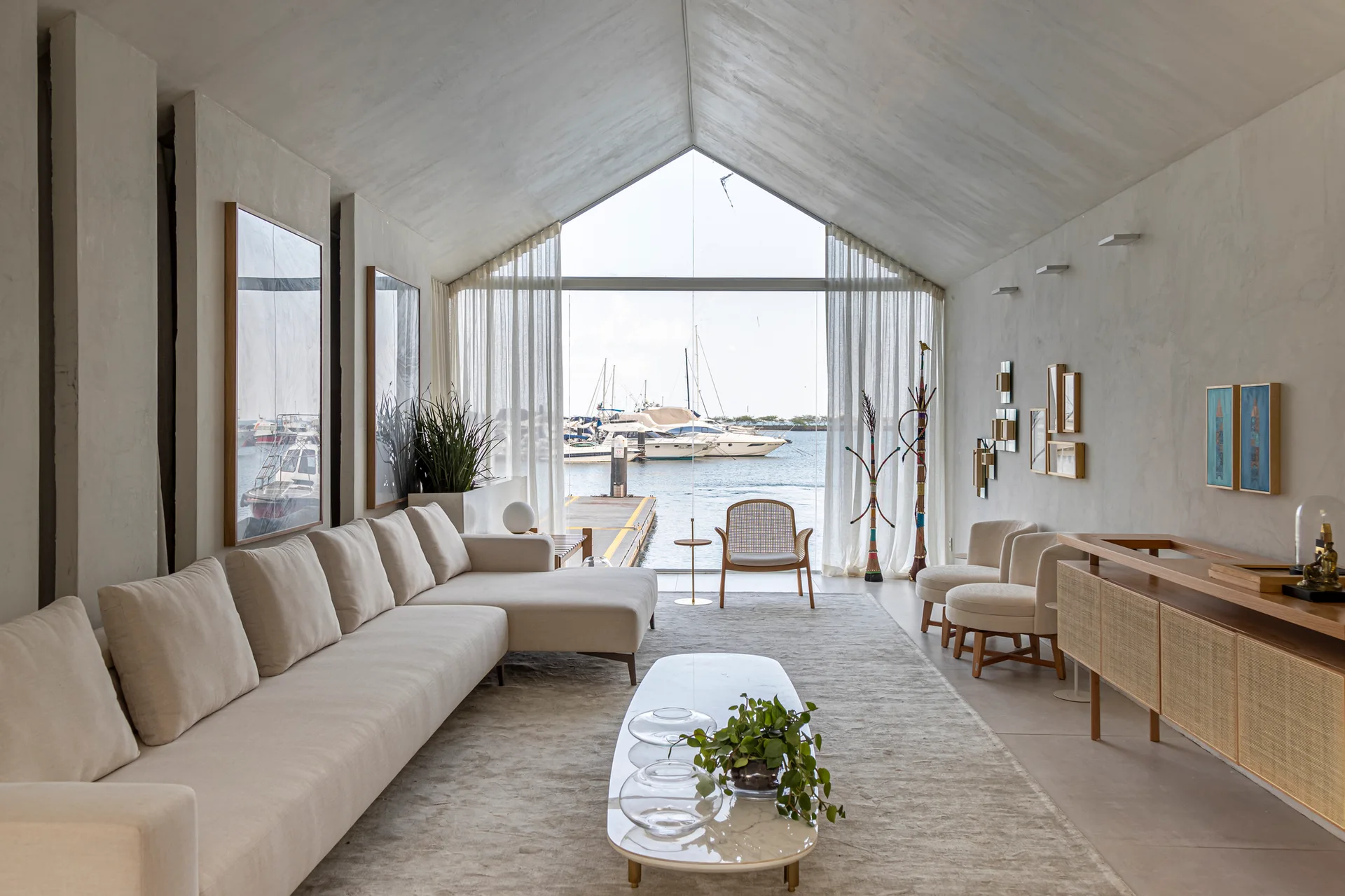 A light-filled living room featuring a long cream sectional sofa, a marble coffee table, and a large gable window looking out onto a marina with boats.