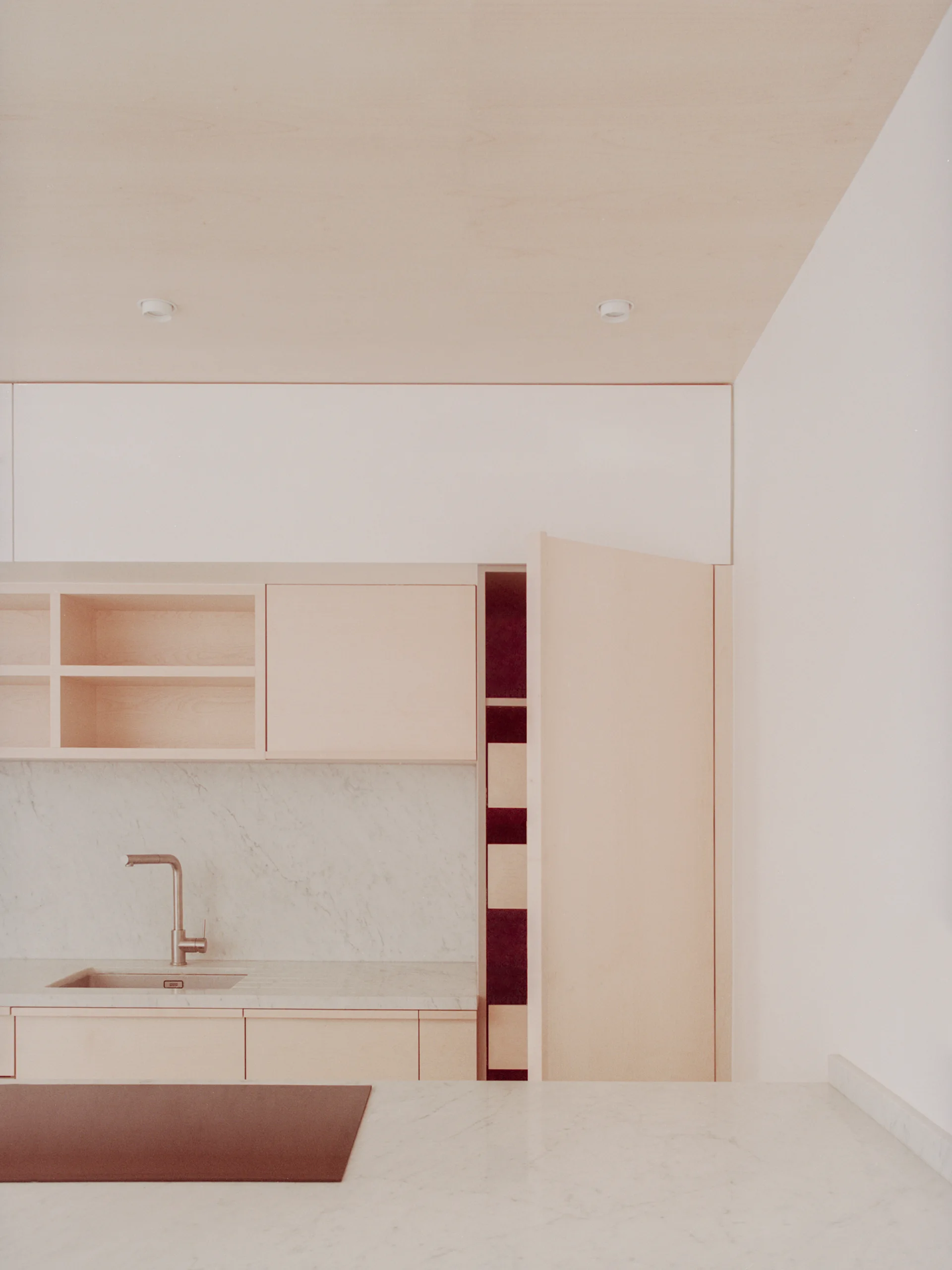 A minimalist kitchen features pale wood cabinets, a marble backsplash, and a tall open pantry cabinet with burgundy shelving.
