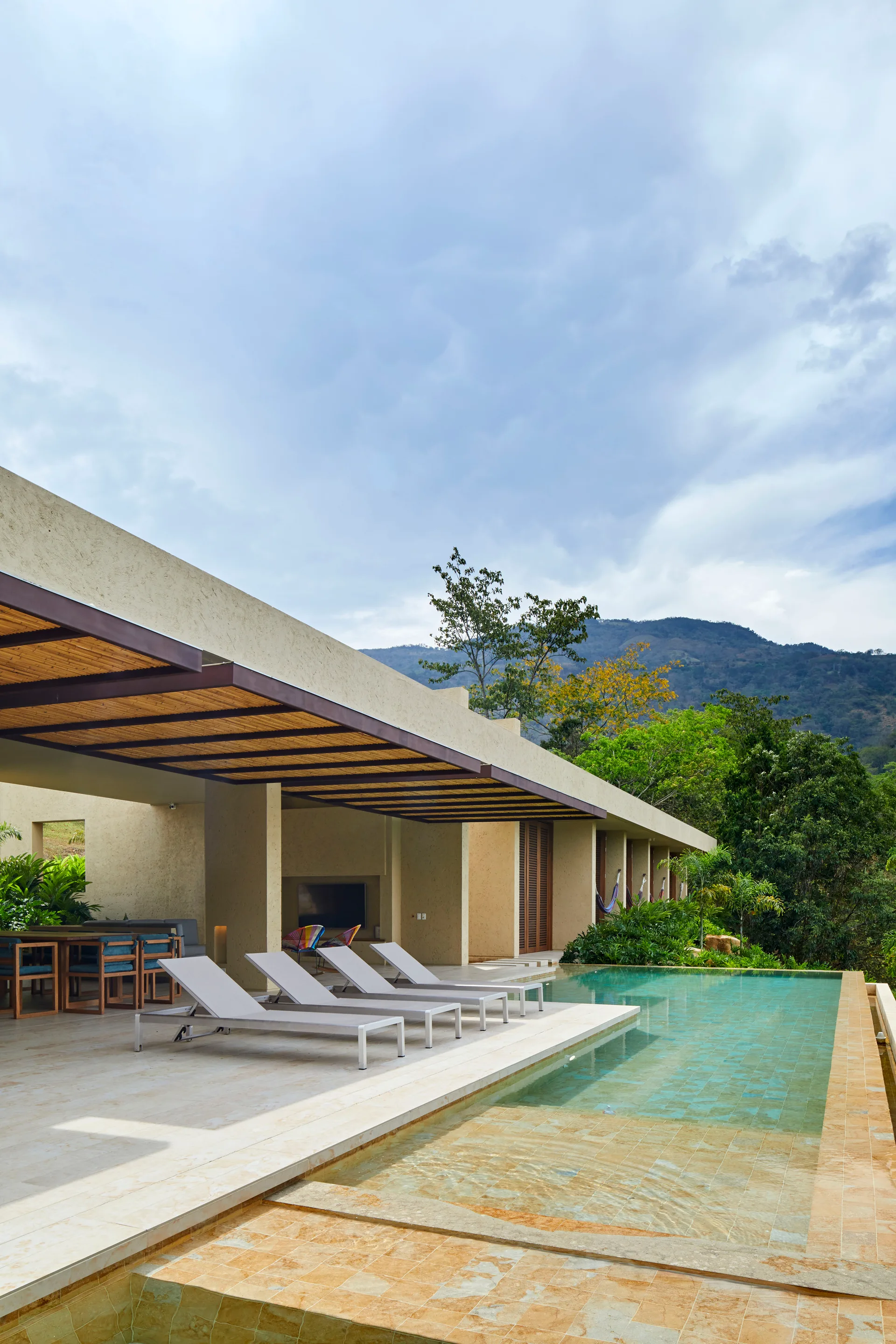 Four white lounge chairs sit beside a rectangular swimming pool on a beige stone patio under a concrete pergola with a slatted bamboo ceiling.