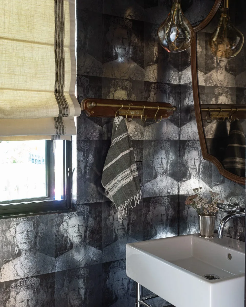 A powder room features dark, repeating portrait-motif wallpaper, a white ceramic sink, a striped roman shade over a window, and a wooden towel rack.