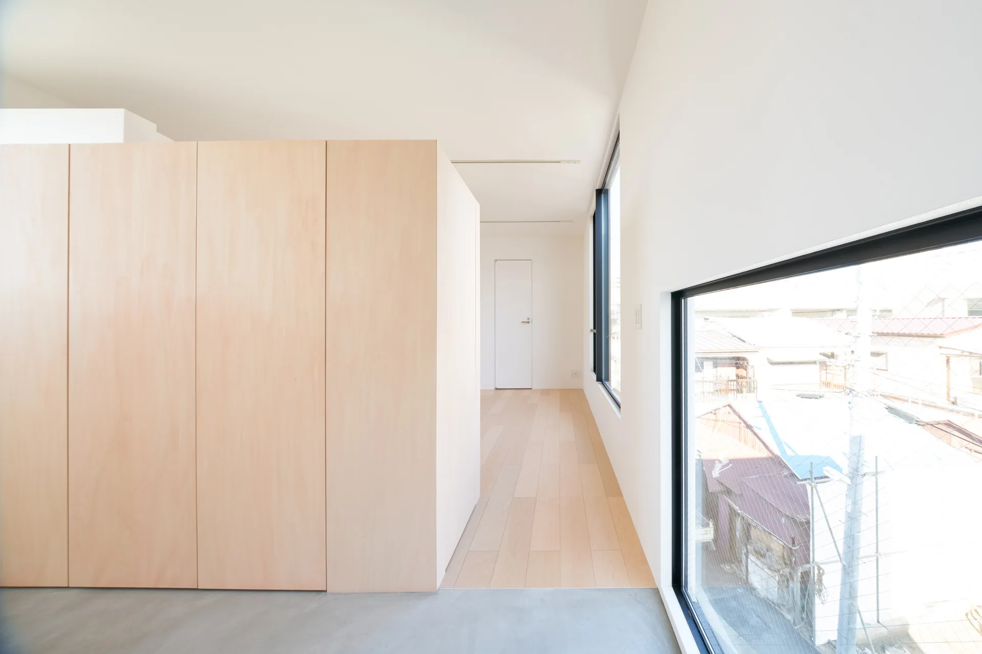 A minimalist hallway with a large light wood cabinet, white walls, and a floor transition from concrete to light wood planks by a low window.