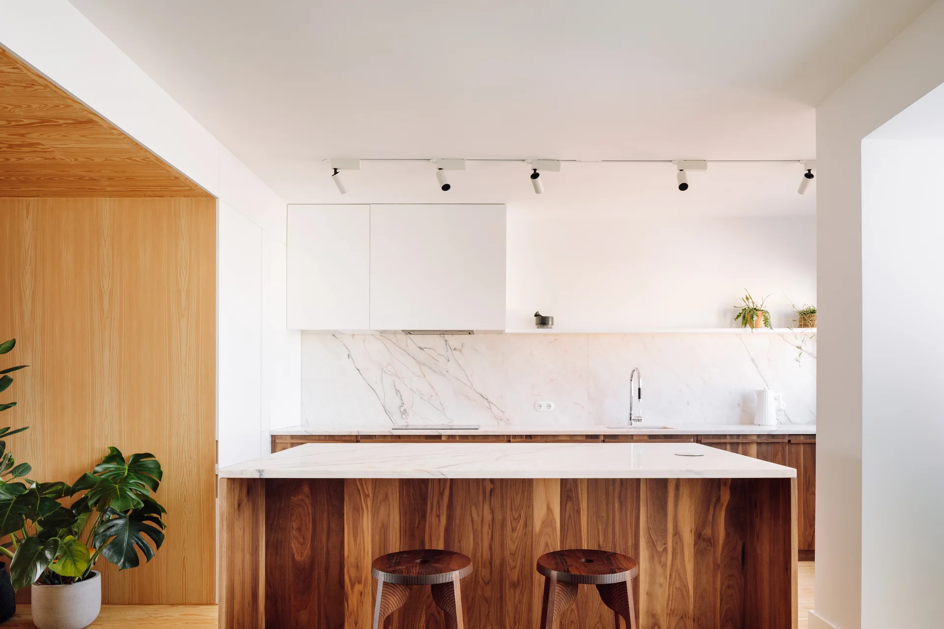 A minimalist kitchen features a wood-grain island with two stools, white upper cabinets, and a white marble backsplash.