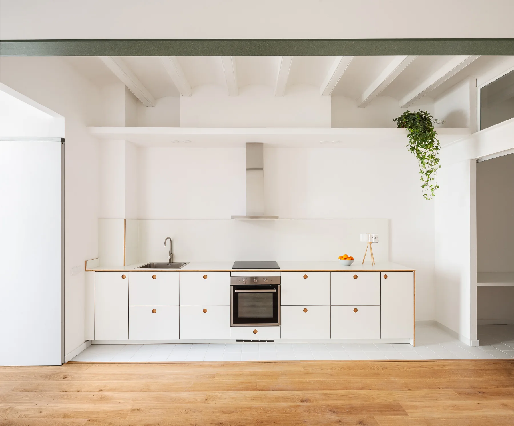 A minimalist white kitchen features flat-front cabinets with circular pulls, a stainless steel range hood, and exposed wooden ceiling beams.