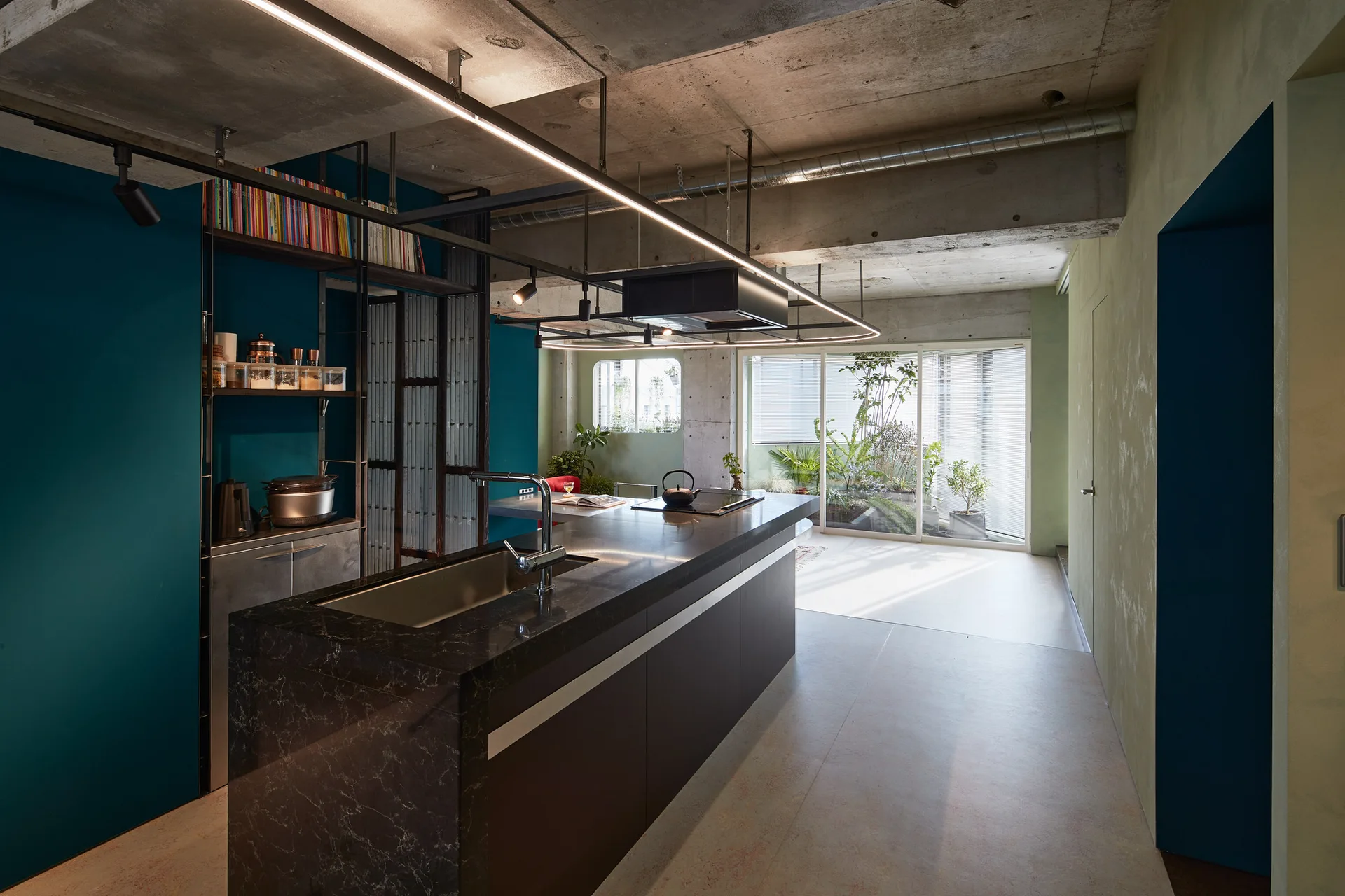 A modern kitchen featuring a dark stone-topped island, a raw concrete ceiling, a suspended linear track light, and teal and sage green walls.