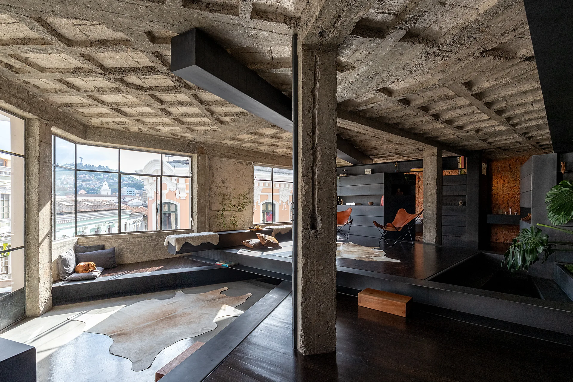An industrial loft living space featuring an exposed concrete waffle ceiling, structural columns, large windows, and a multi-level dark wood floor.