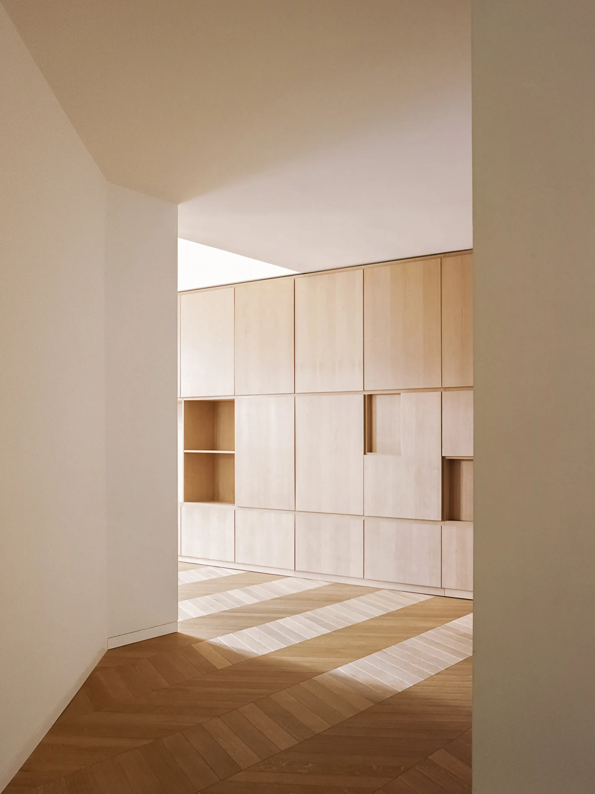 A minimalist hallway features floor-to-ceiling light wood cabinetry with built-in niches and herringbone patterned wood floors.