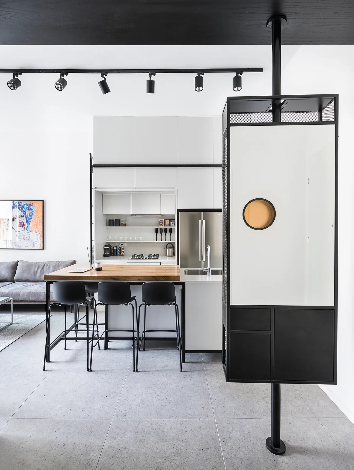 A modern open kitchen features a wood-topped island with black stools, white minimalist cabinets, and a black pole-mounted room divider on grey tile floors.