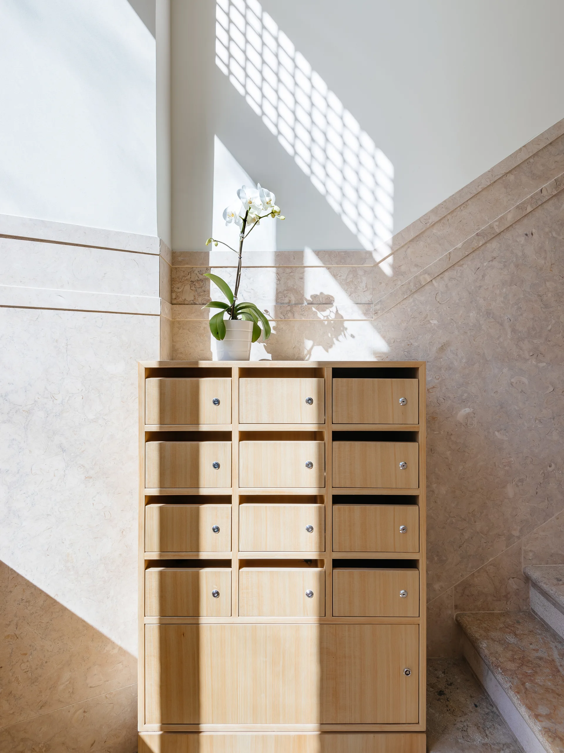 A light wood storage cabinet with small cubby doors sits against a beige marble-clad wall beside stone stairs.