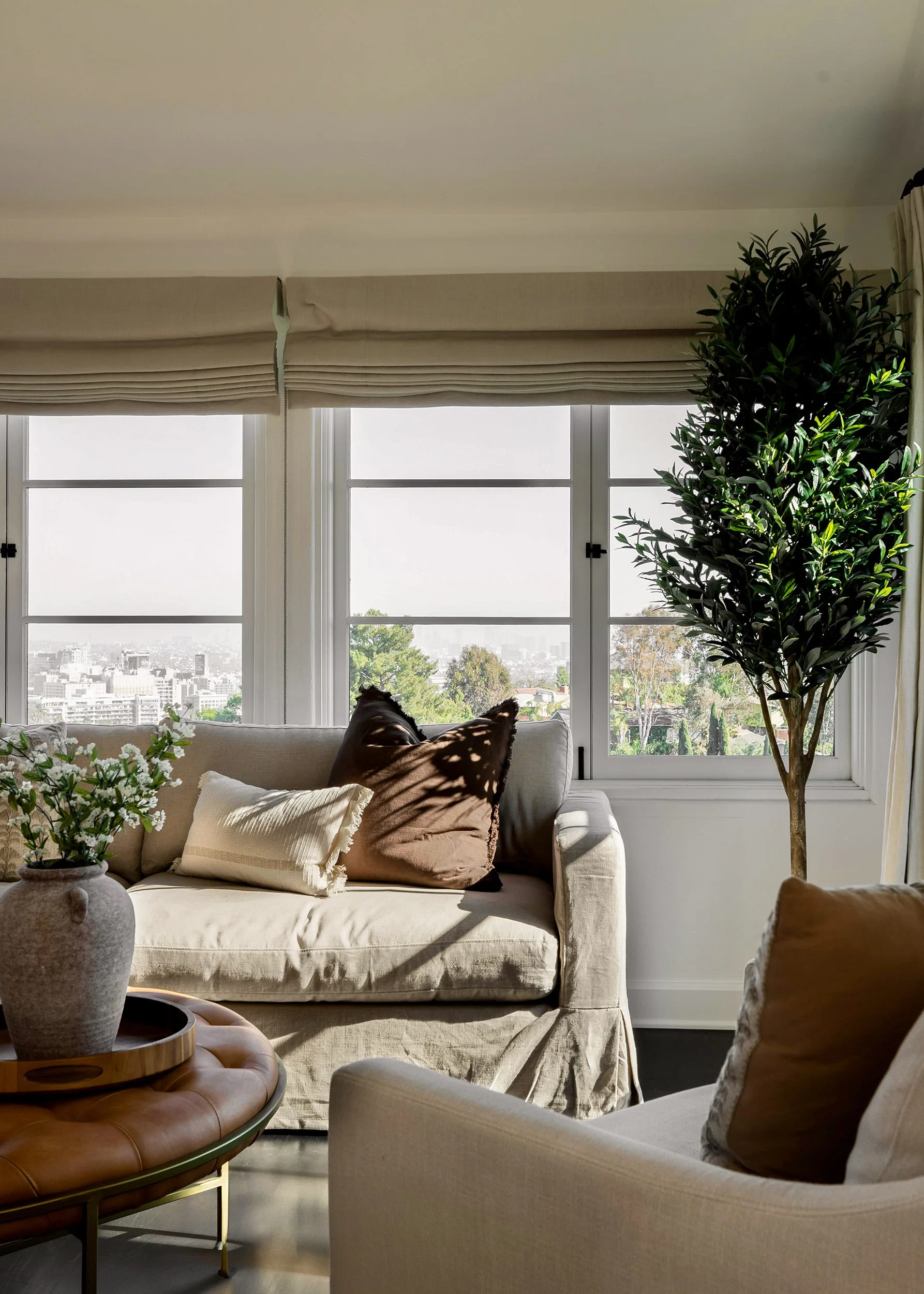 A sunlit living room features a beige slipcovered sofa in front of large windows with fabric Roman shades next to a tall potted olive tree.