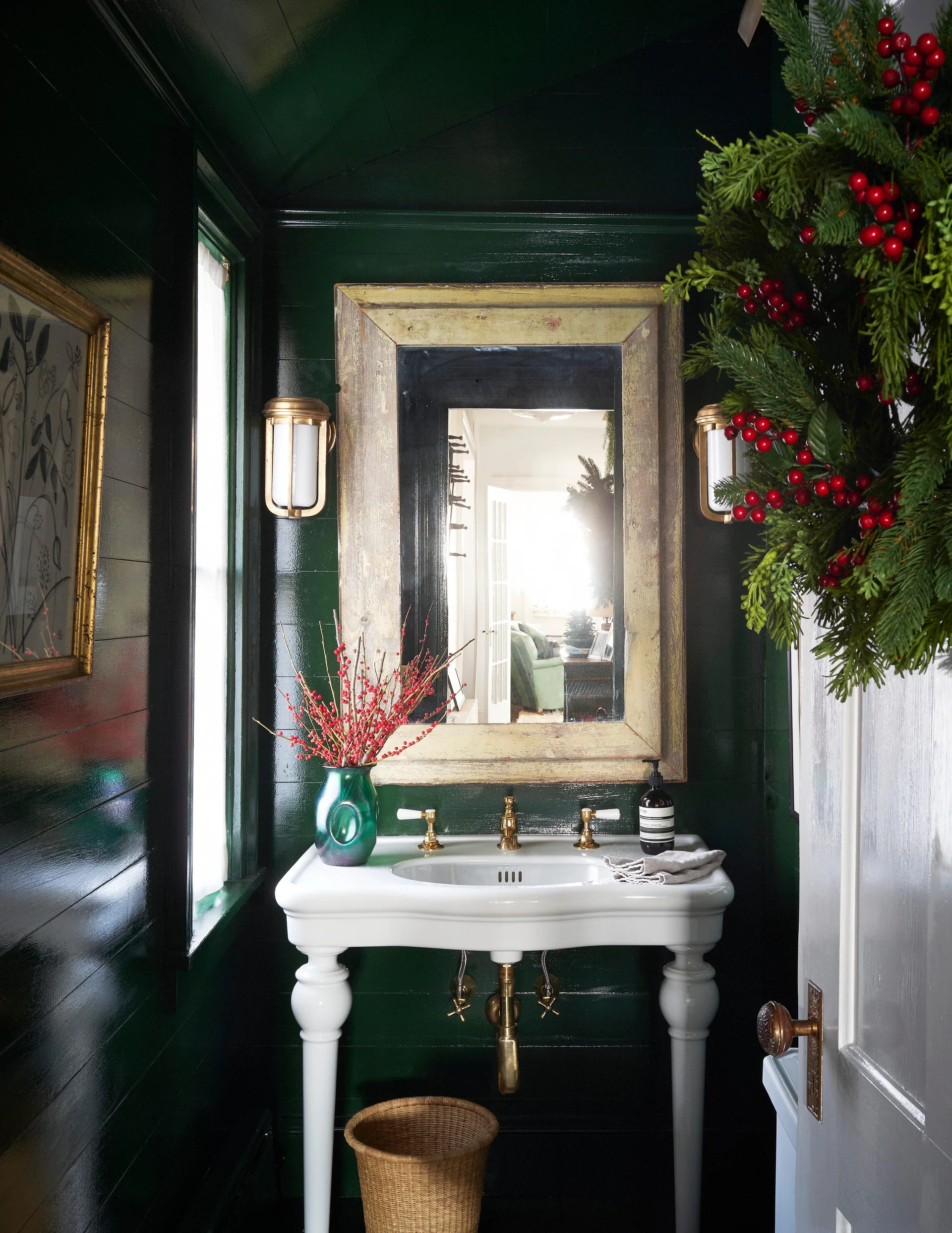A small powder room features glossy forest green wood paneled walls, a white console sink with brass fixtures, and a weathered wooden mirror.