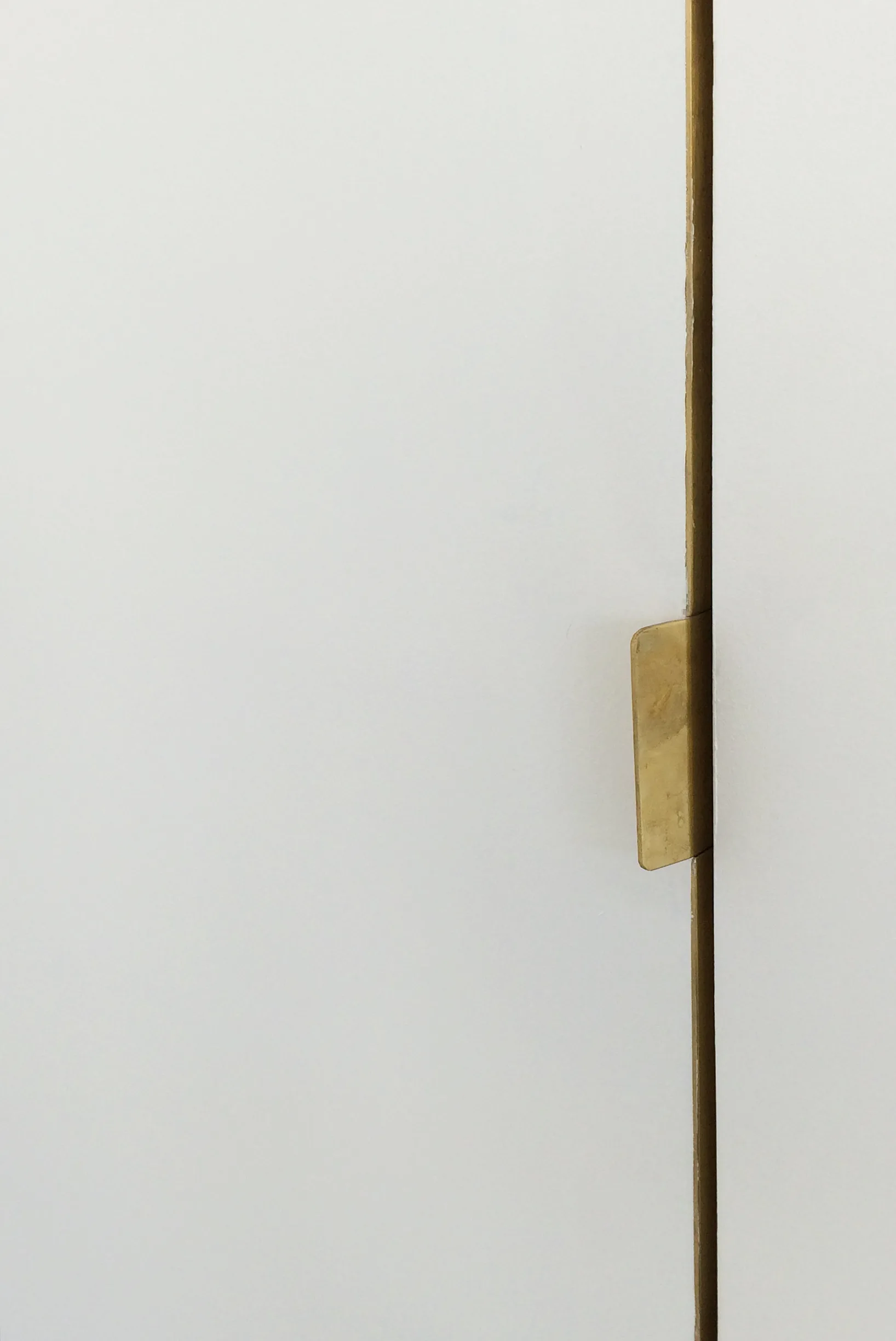 A close-up of a matte white cabinet door with a small, flat brass edge pull handle integrated along the vertical seam.