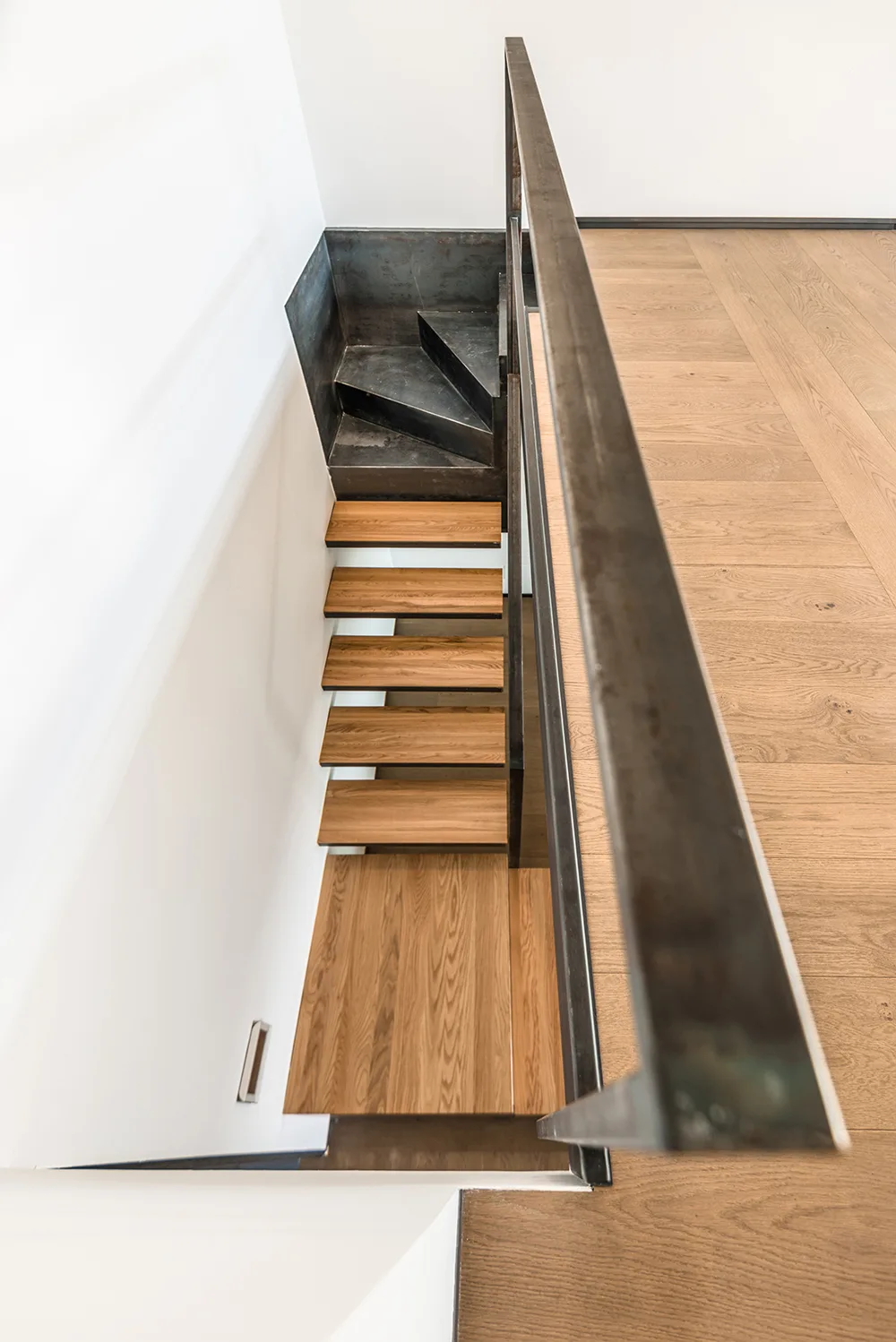 A high-angle view looking down a minimalist staircase with natural wood treads, a dark metal railing, and smooth white walls.