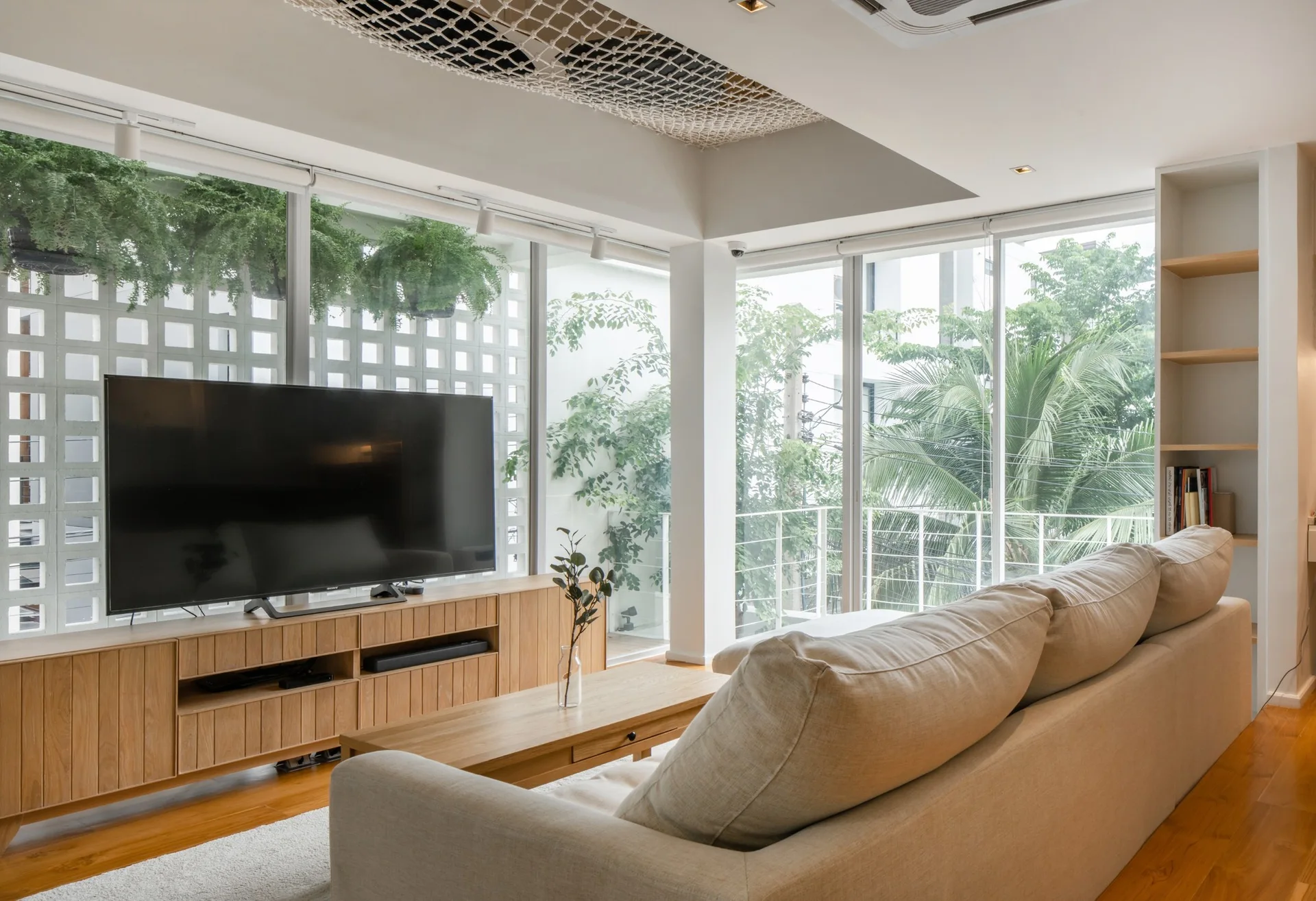 A bright living room features a beige sofa, a light wood media console with a television, and large windows looking out at green plants.