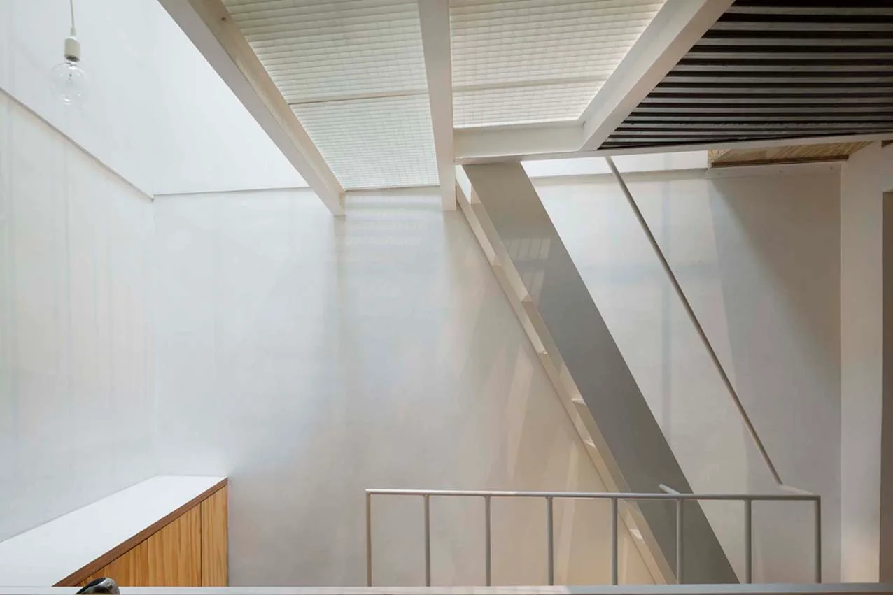 A bright minimalist stairwell features white painted walls, a metal railing, and a translucent gridded ceiling beneath an overhead skylight.