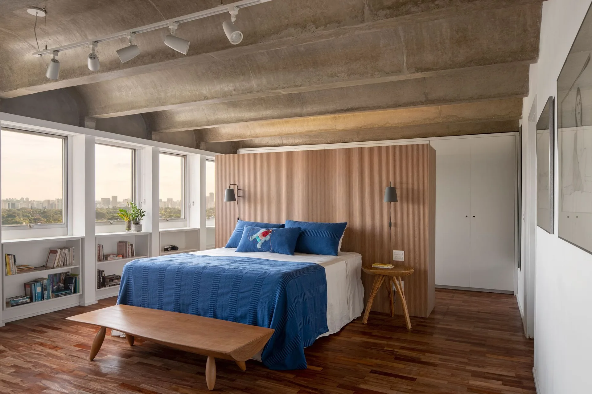 A contemporary bedroom featuring a bed with blue bedding set against a fluted wood partition beneath an exposed concrete ceiling.