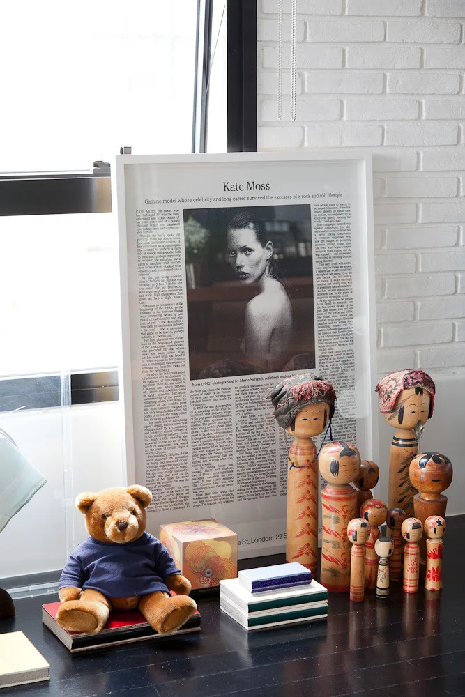 A collection of wooden kokeshi dolls, a teddy bear, and stacked books arranged on a dark shelf in front of a leaning framed print and a white brick wall.