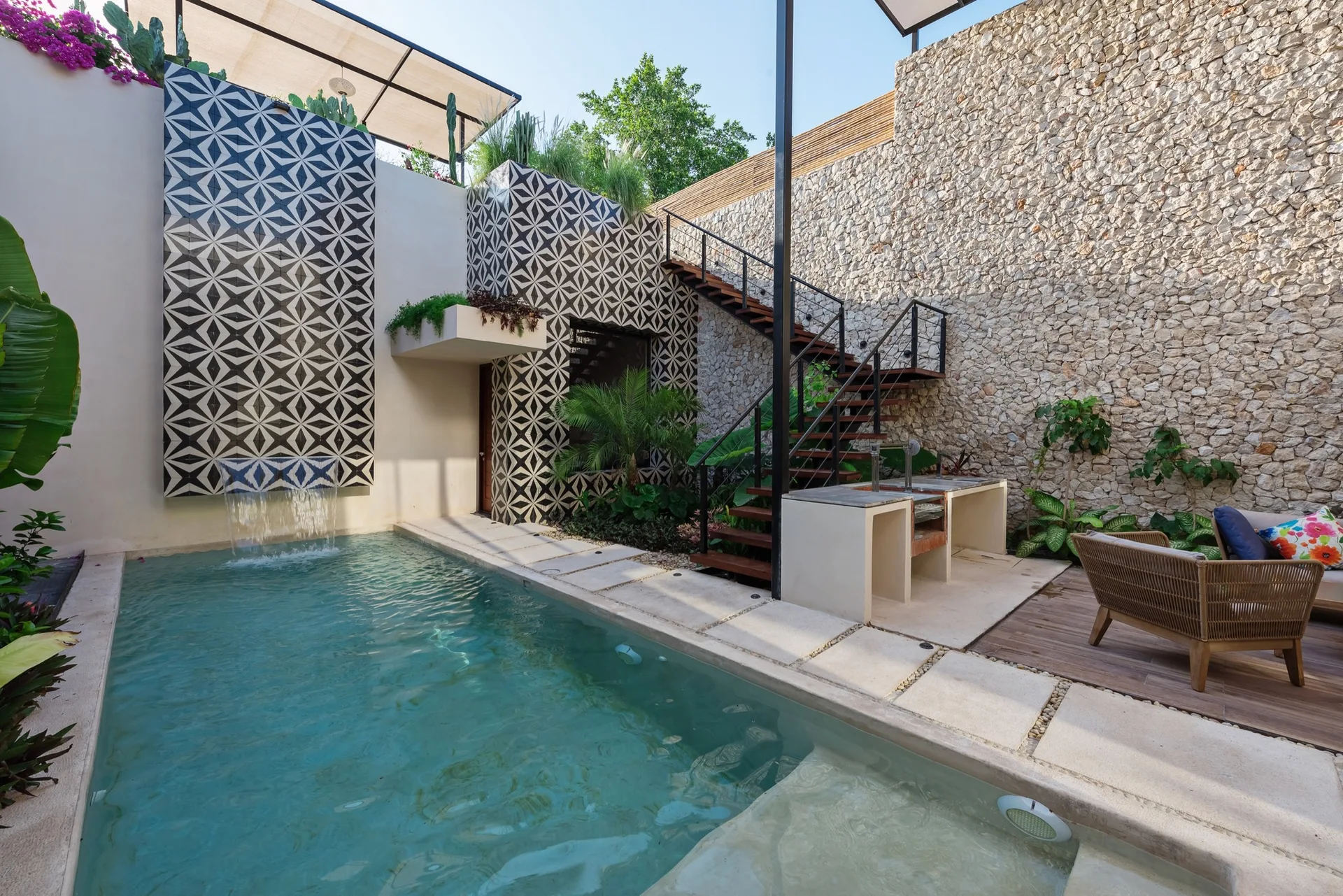 A backyard swimming pool features a geometric black and white tile waterfall, a natural stone wall, a wooden staircase, and a concrete outdoor kitchen.