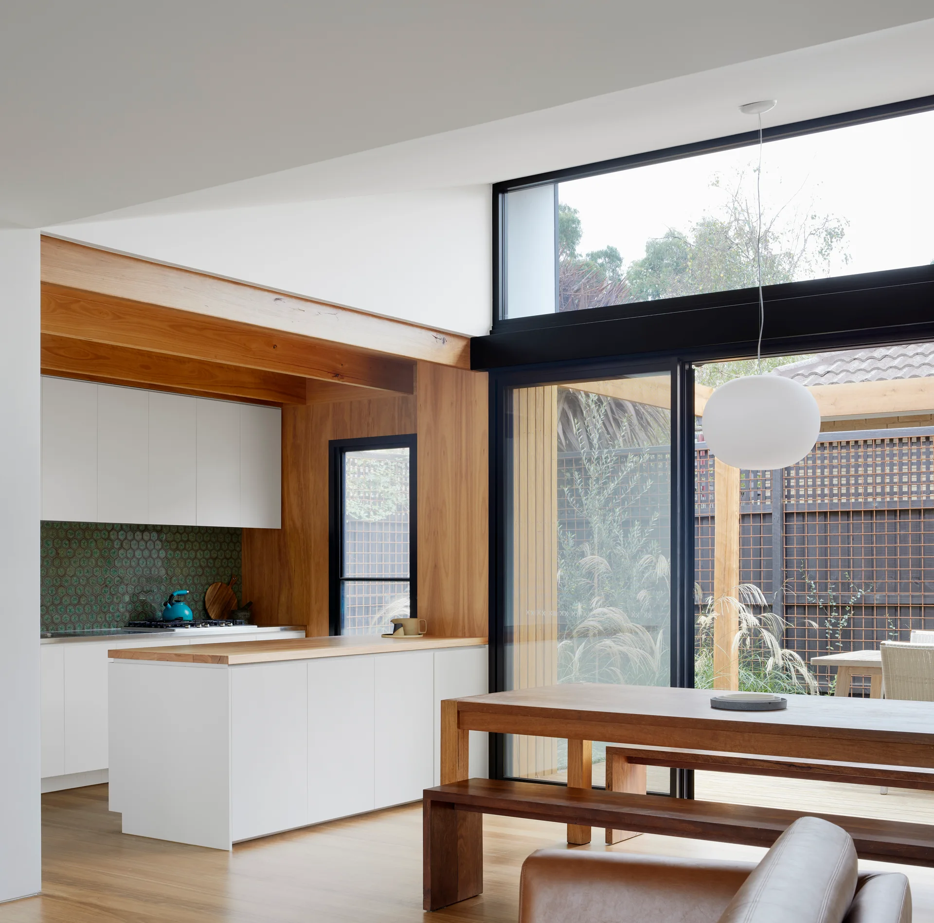 A modern kitchen and dining area featuring white cabinets framed in wood paneling, a green hex tile backsplash, and a wooden table.