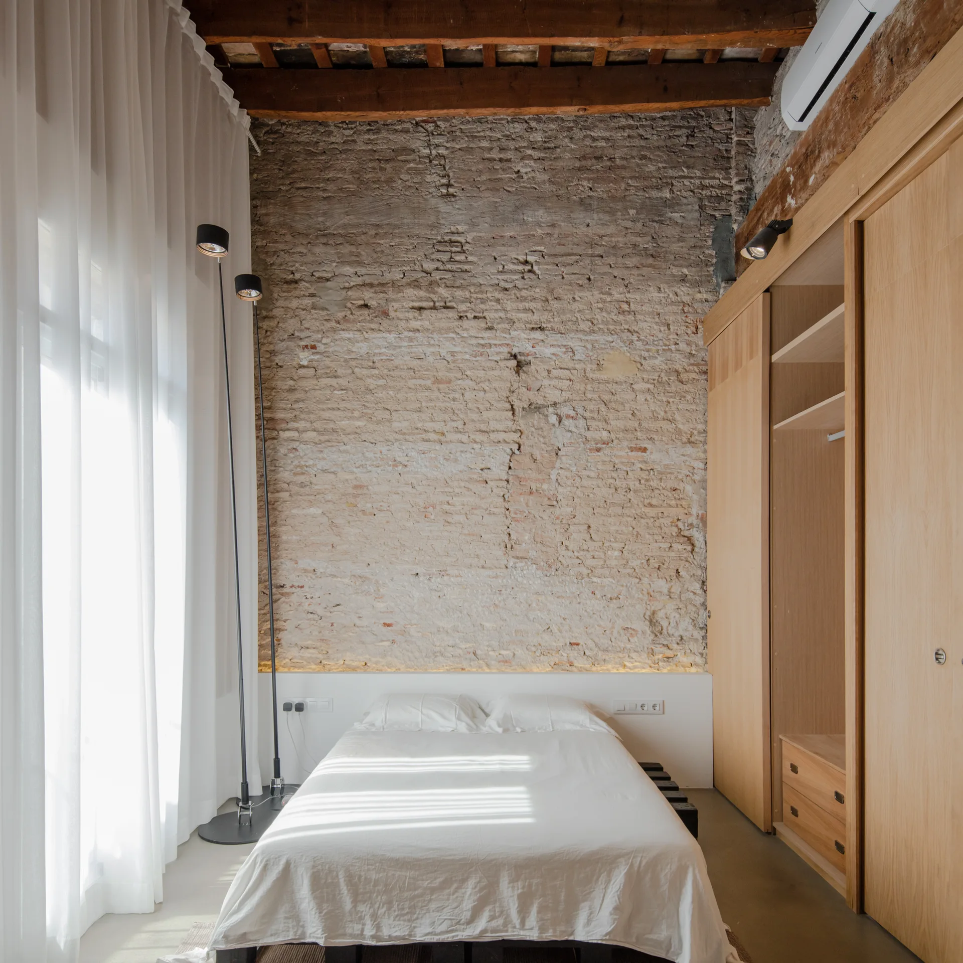 A minimalist bedroom featuring an exposed brick wall behind a white bed, a built-in timber wardrobe, and sheer white curtains.