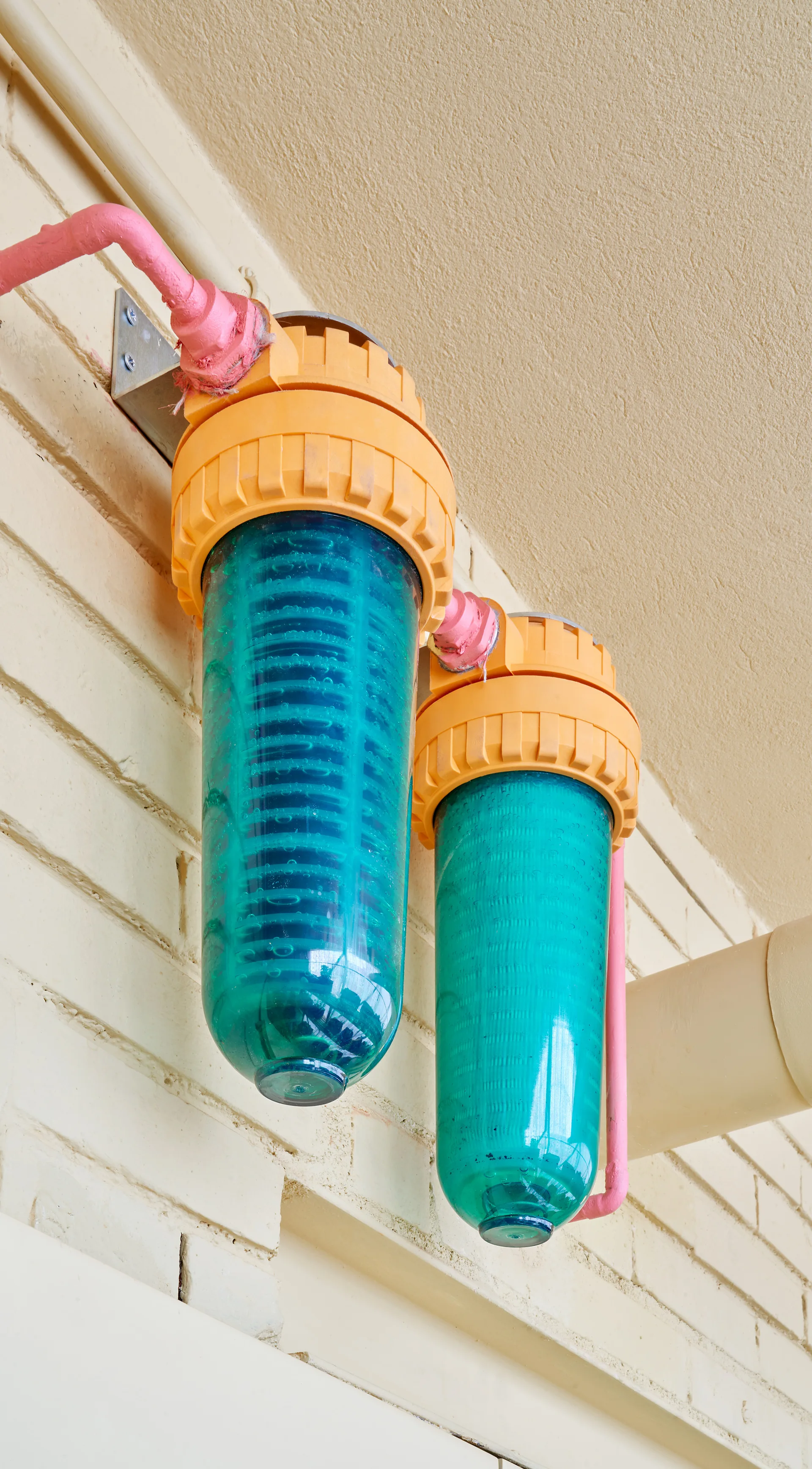 Two translucent turquoise water filters with orange caps and pink piping are mounted on a painted beige brick wall.