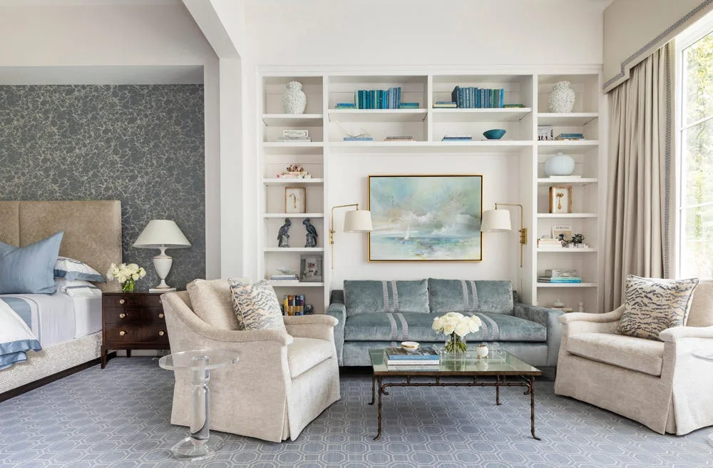 A bedroom seating area features a blue velvet sofa tucked into a white built-in shelving unit, flanked by two cream armchairs on a patterned grey carpet.