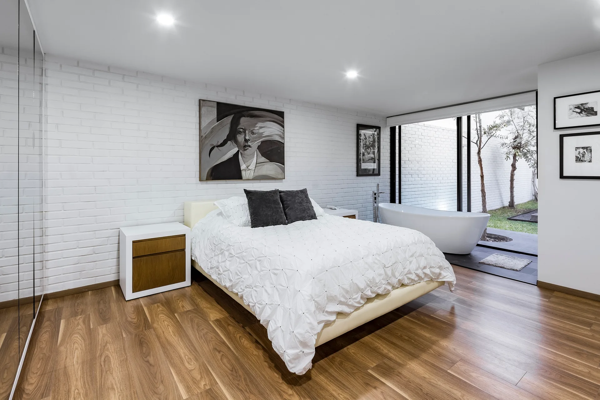 A modern bedroom features a bed with white textured bedding, a white brick accent wall, wood flooring, and a freestanding bathtub near a courtyard window.