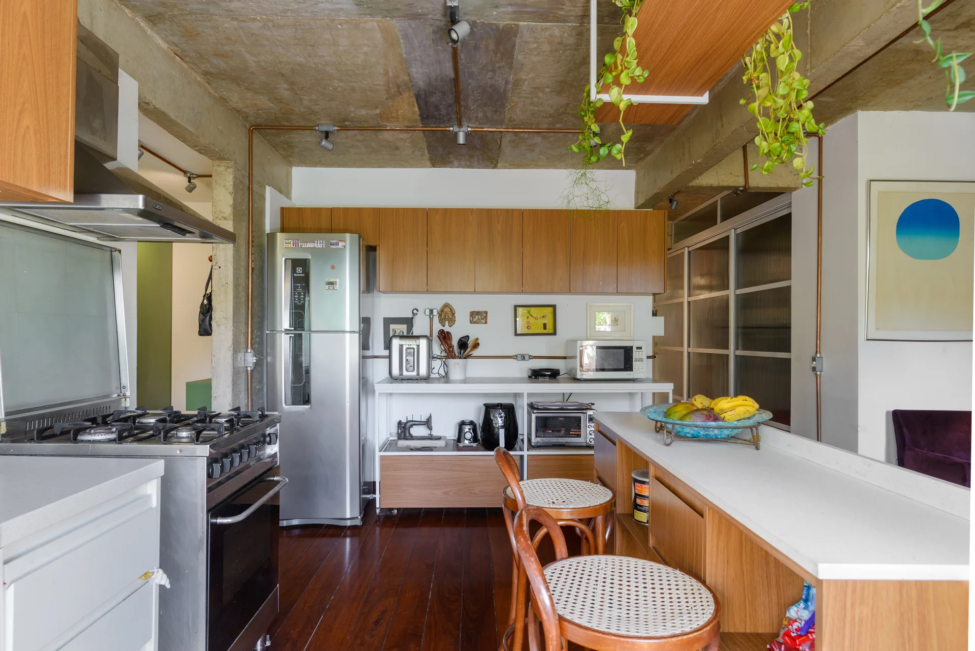 An industrial kitchen with an exposed concrete ceiling, copper pipes, warm wood cabinets, and a kitchen island with cane seat bar stools.