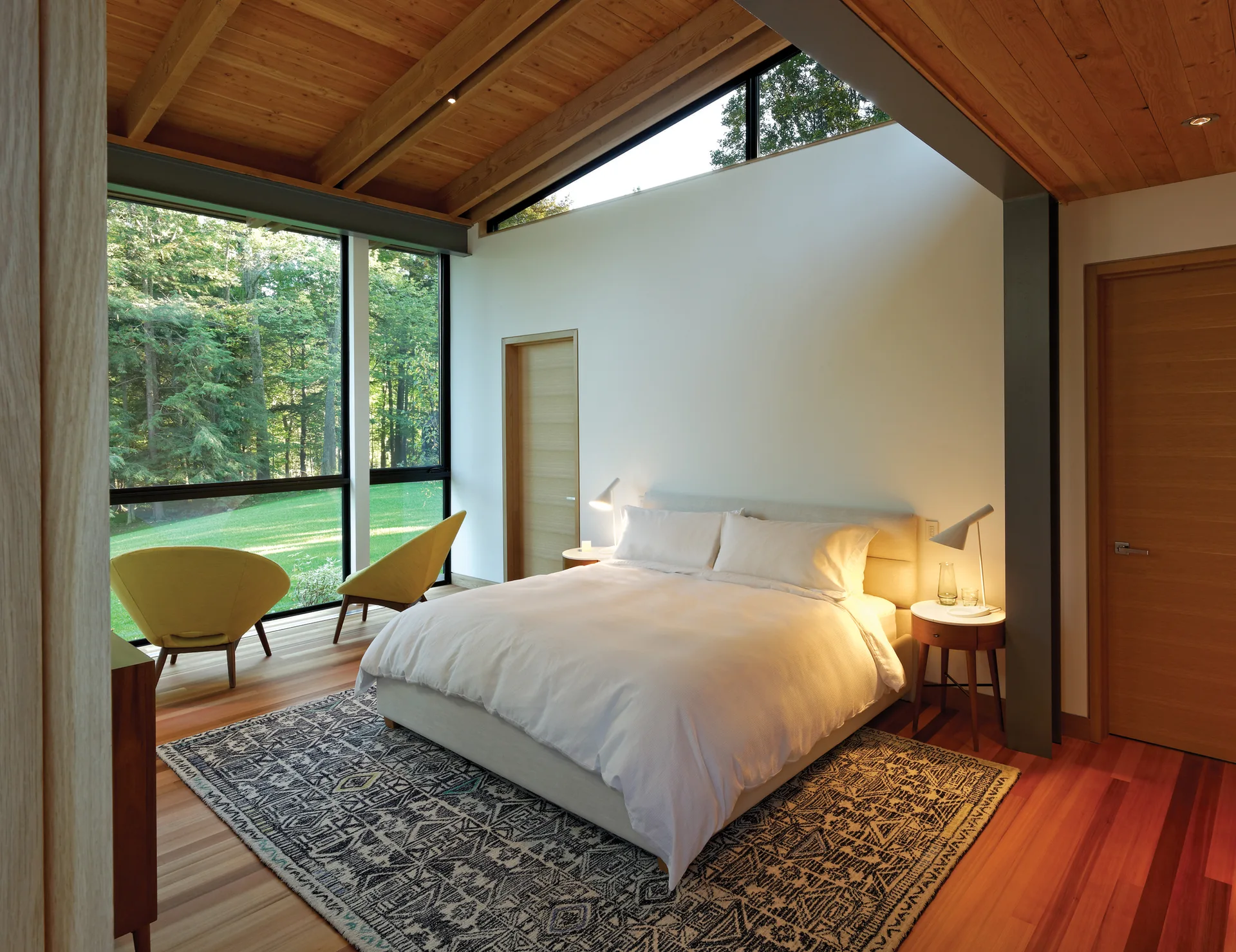 A modern bedroom features a pitched wooden ceiling, floor-to-ceiling windows looking out to a forest, a neutral bed, and two yellow accent chairs.