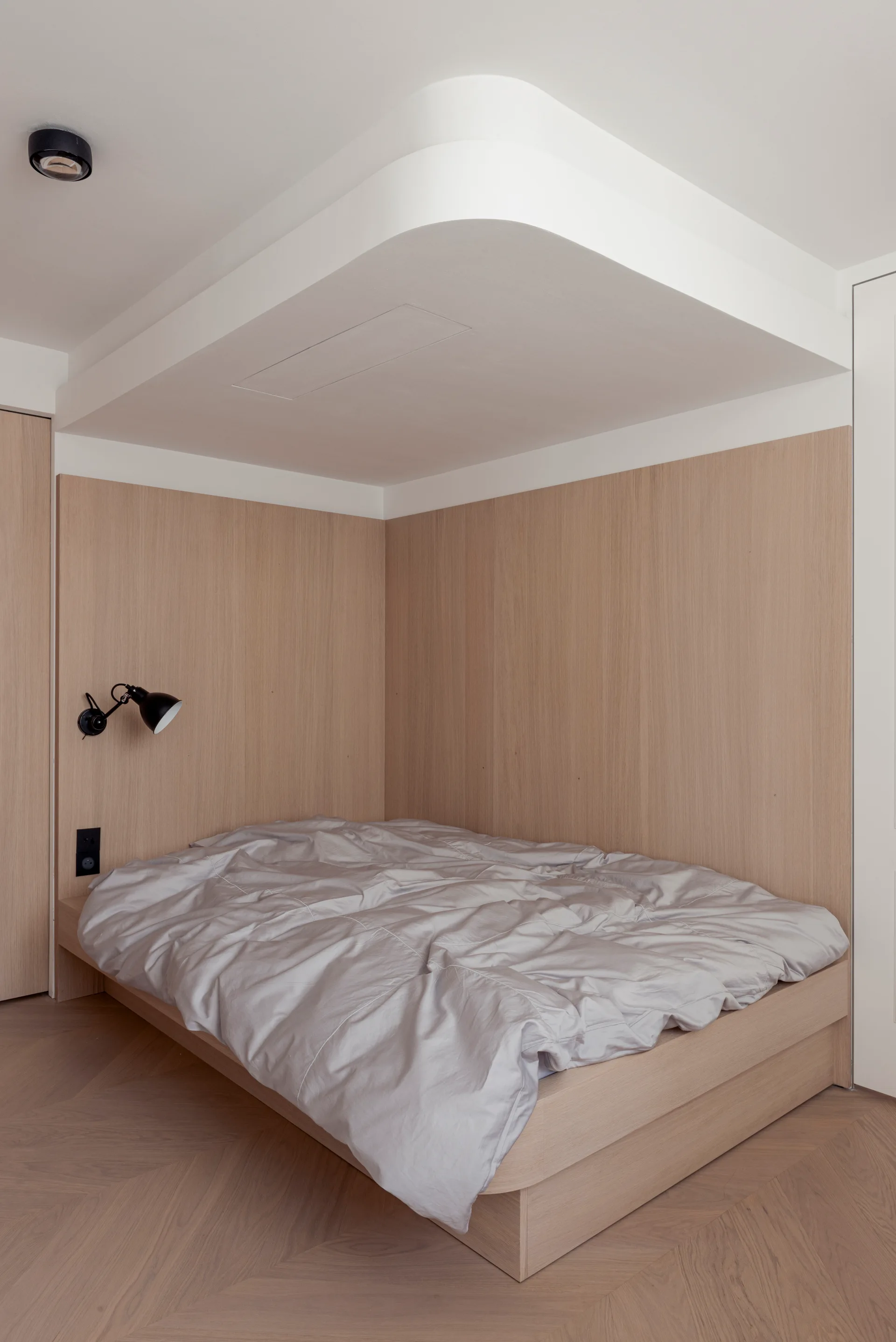 A minimalist bedroom features a built-in light wood platform bed matching the wall paneling under a white curved dropped ceiling.