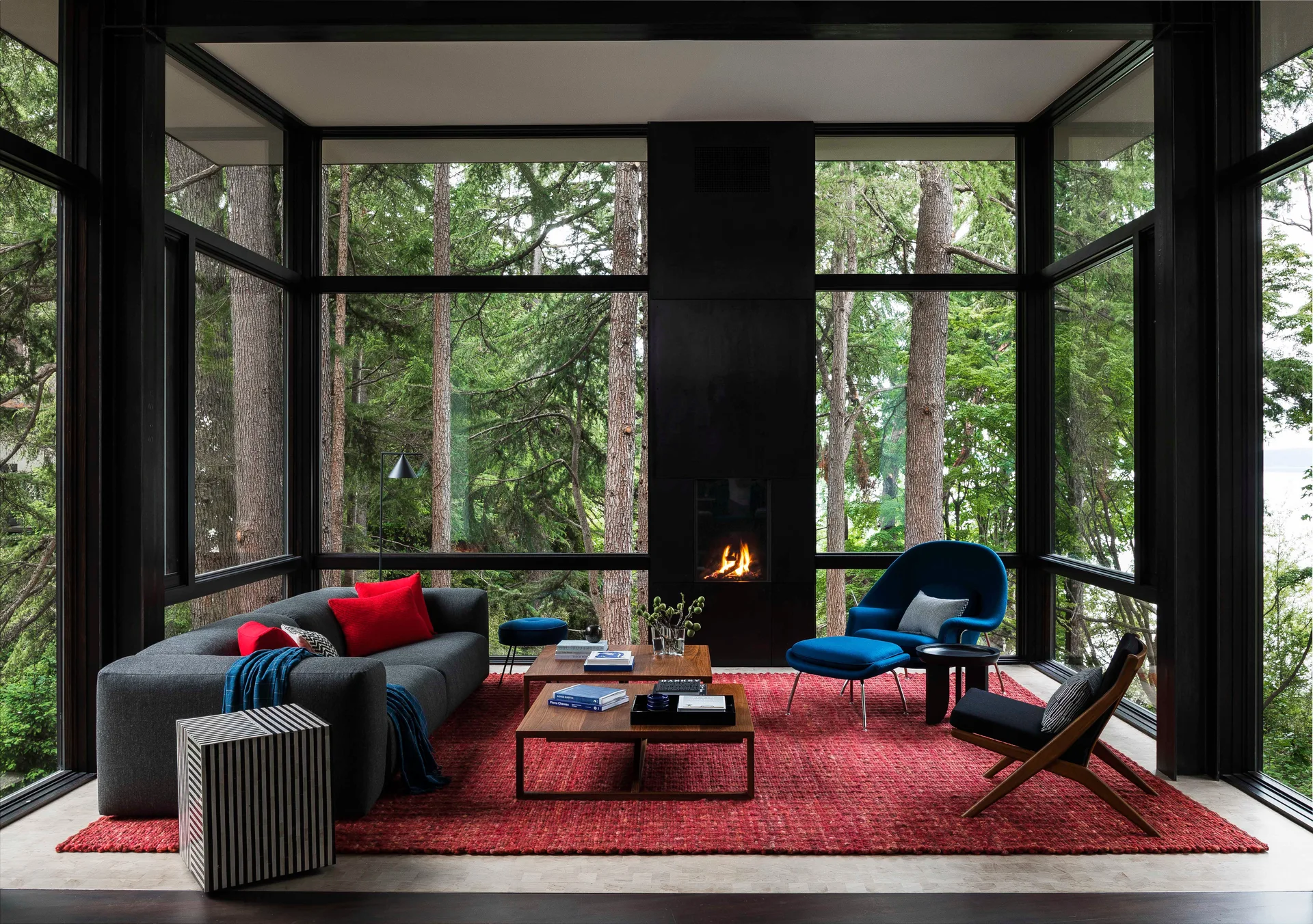 A modern living room featuring tall glass walls with forest views, a black fireplace, a grey sofa, a blue armchair, and a red rug.