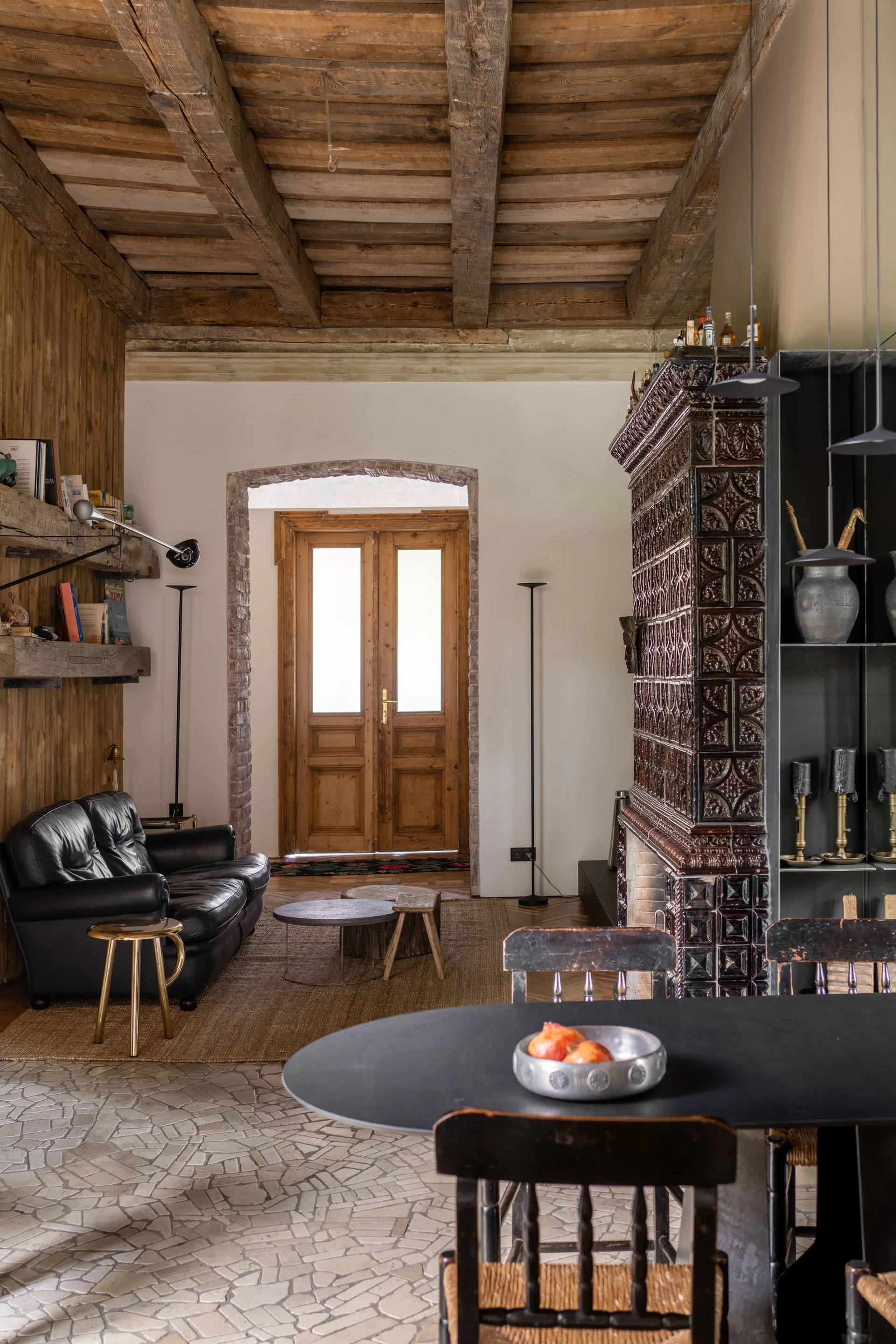A rustic living room features a black leather sofa, a large ornate ceramic tiled stove, exposed wooden ceiling beams, and stone tile flooring.
