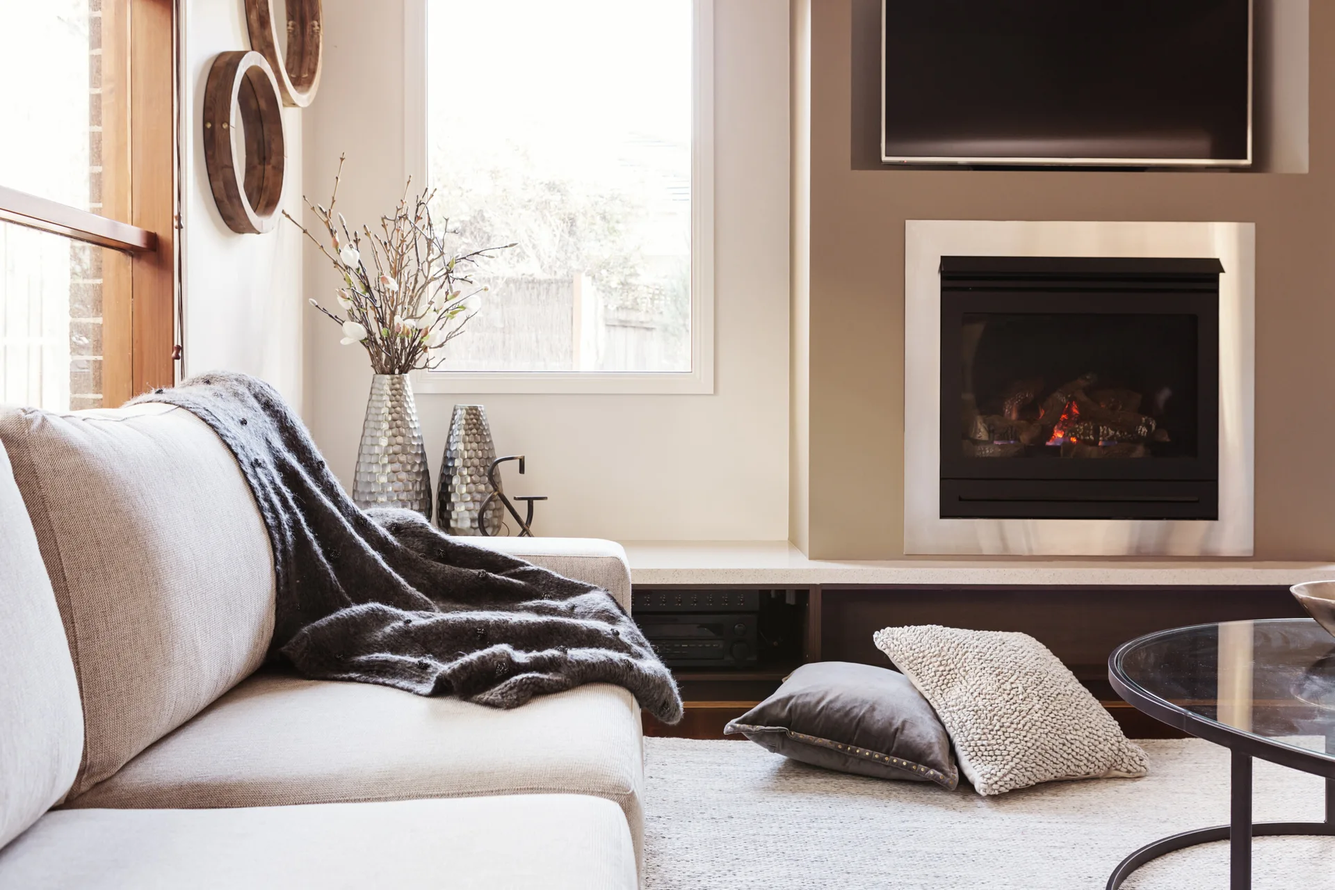 A light beige sofa sits near a window next to a fireplace with a brushed metal surround and mounted television.