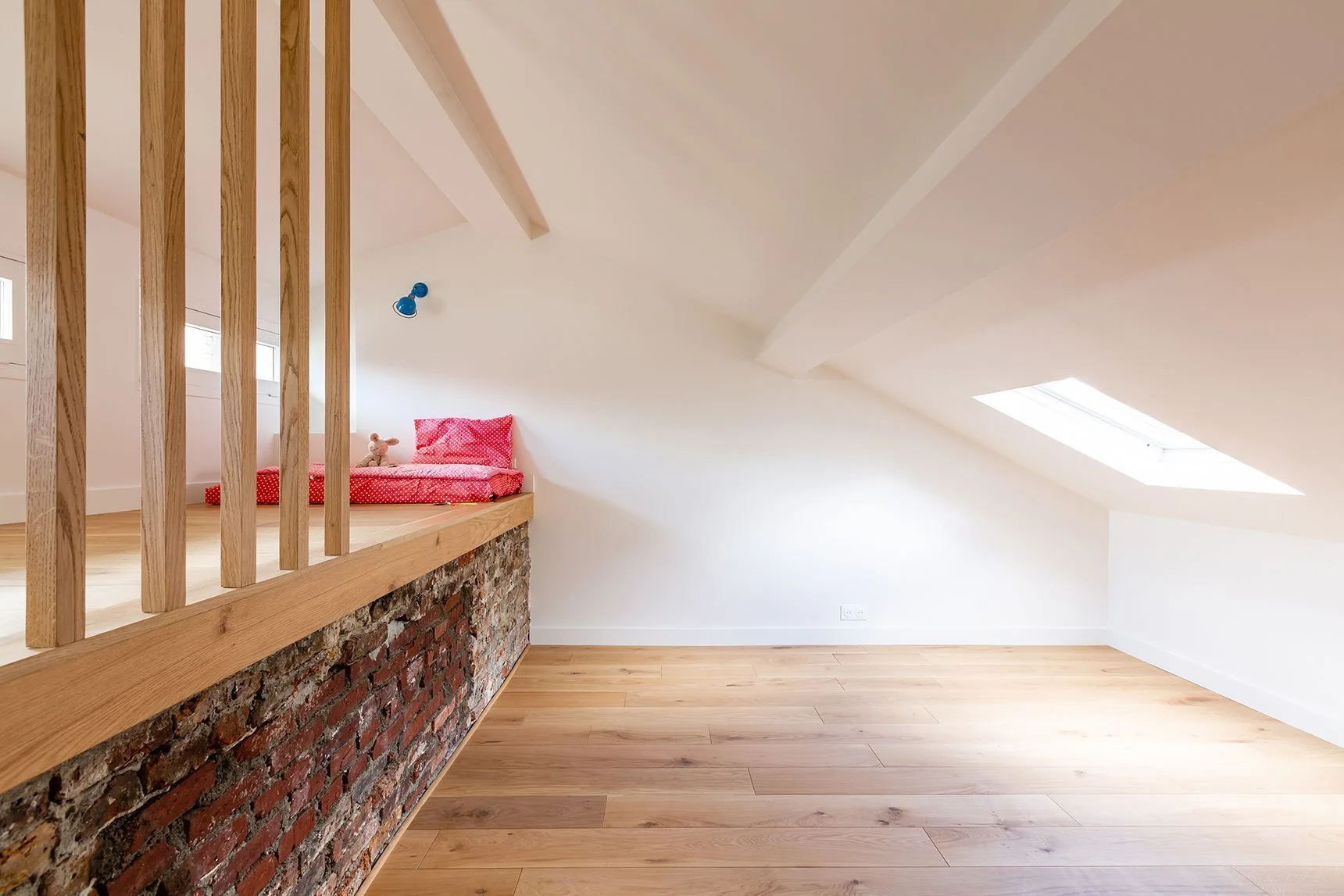 An attic kids room with a wooden loft platform built over an exposed brick wall next to white sloped ceilings and wood flooring.
