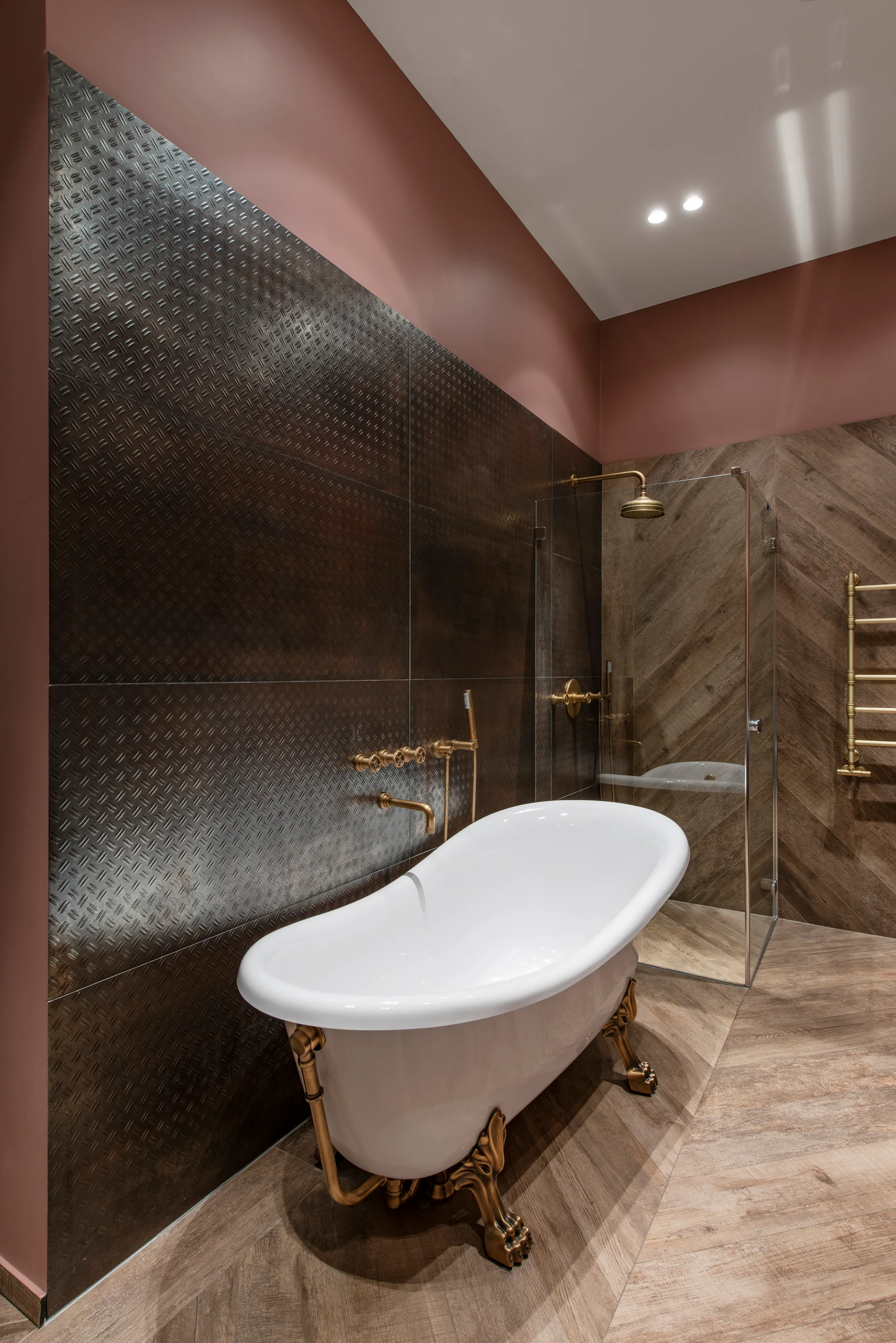 A white clawfoot bathtub with brass legs sits on a herringbone floor next to dark diamond plate wall tiles and a glass-enclosed shower.