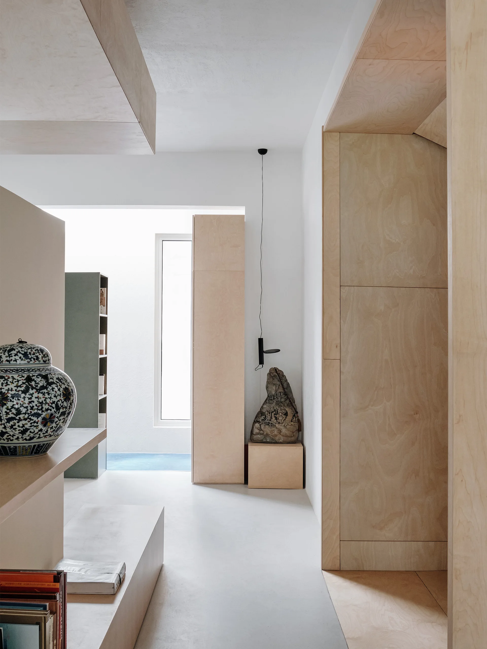 A minimalist hallway features light-colored plywood paneling and built-in cabinets, a smooth white floor, a patterned blue vase, and a distant window.