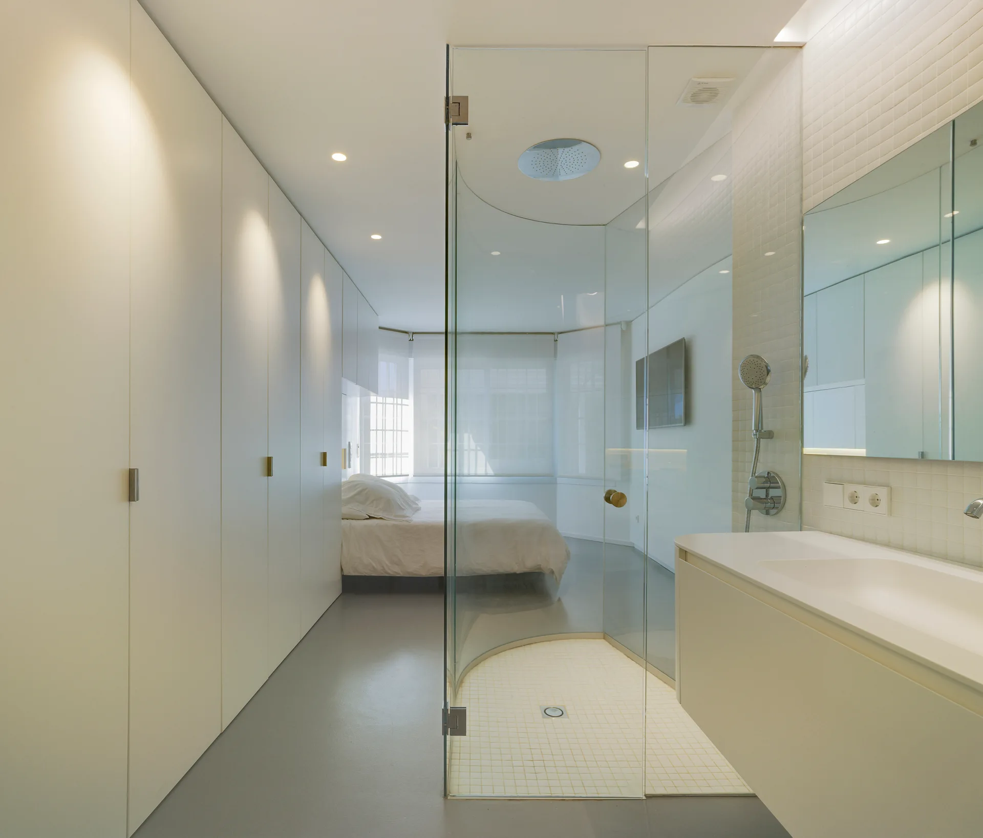 A minimalist bedroom suite featuring a curved glass shower enclosure, a white bathroom vanity with a mirror, and a row of floor-to-ceiling closets.