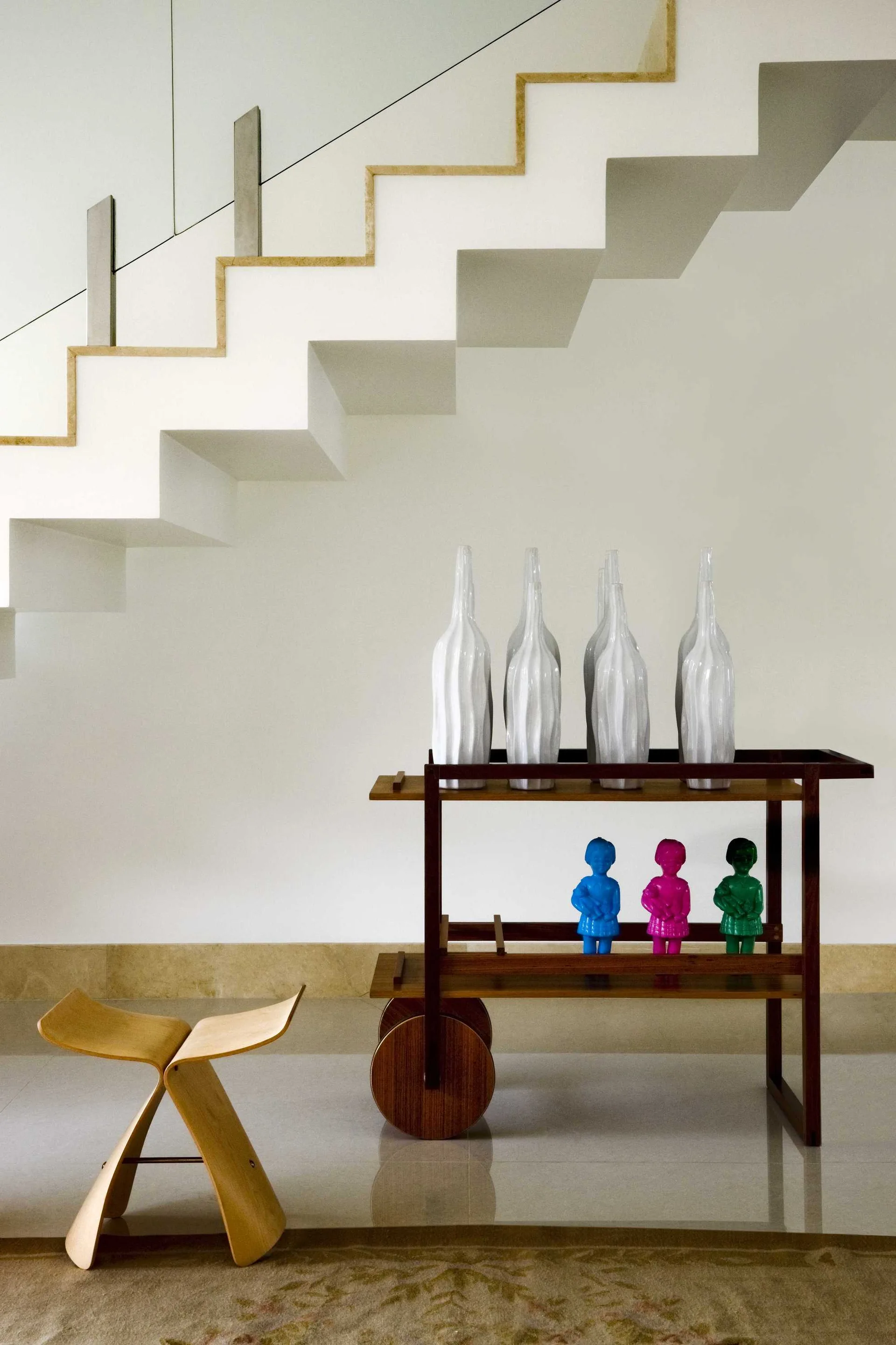 A wooden bar cart styled with tall white vases and colorful figurines stands beneath a modern white staircase next to a low wooden stool.
