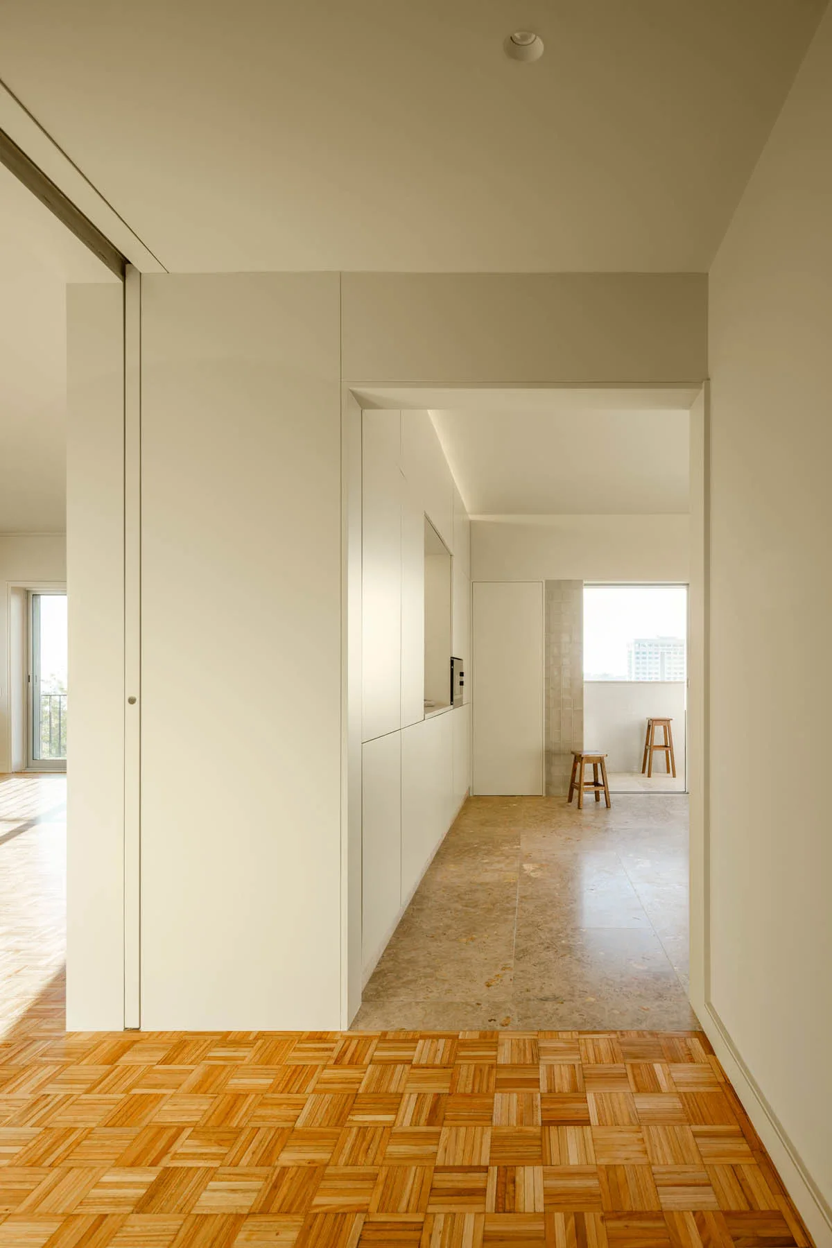 A minimalist hallway with parquet wood flooring transitions into a kitchen area with stone tile floors, white built-in cabinetry, and two wooden stools.