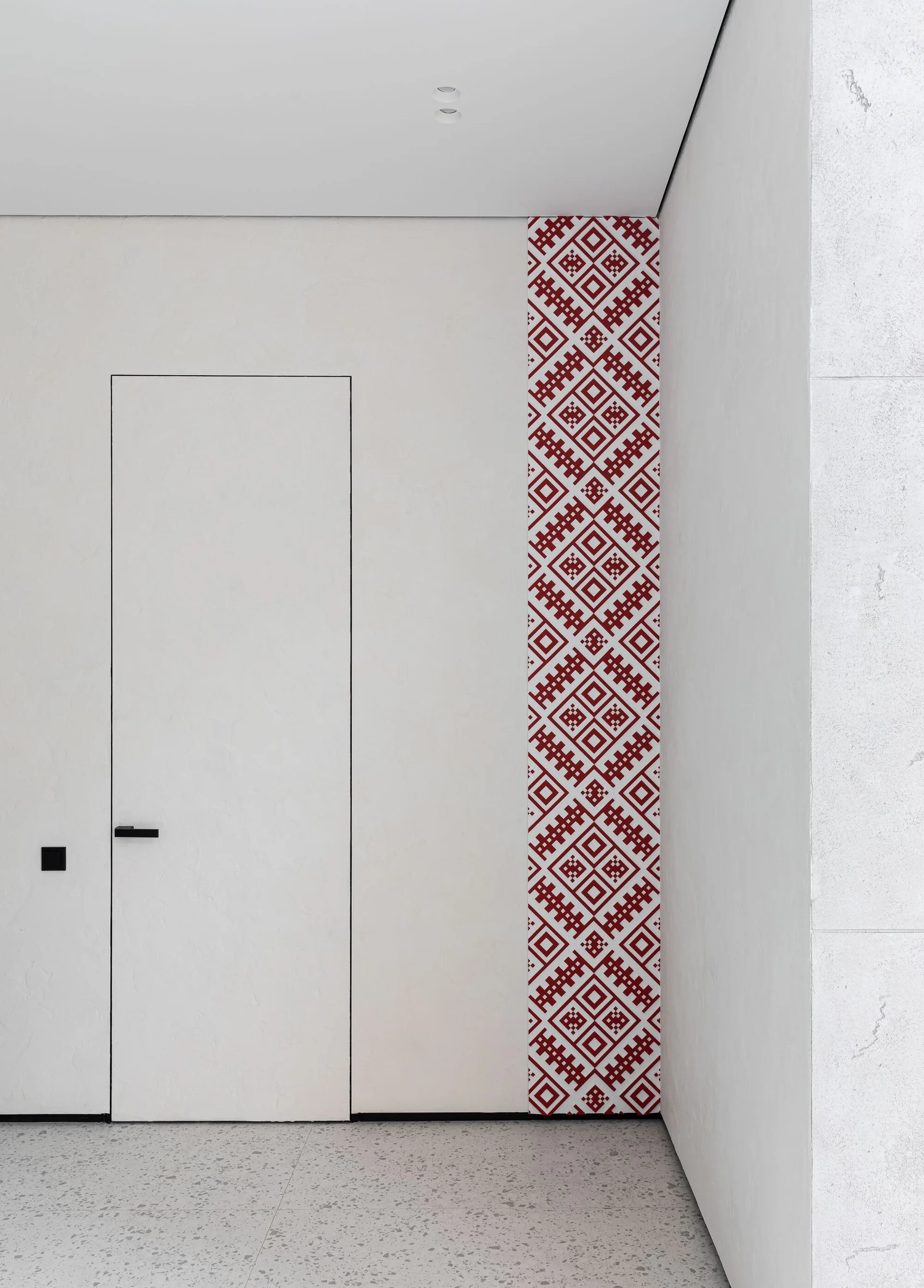 A minimalist white hallway features a flush hidden door with a black handle next to a vertical strip of red geometric pattern and speckled flooring.