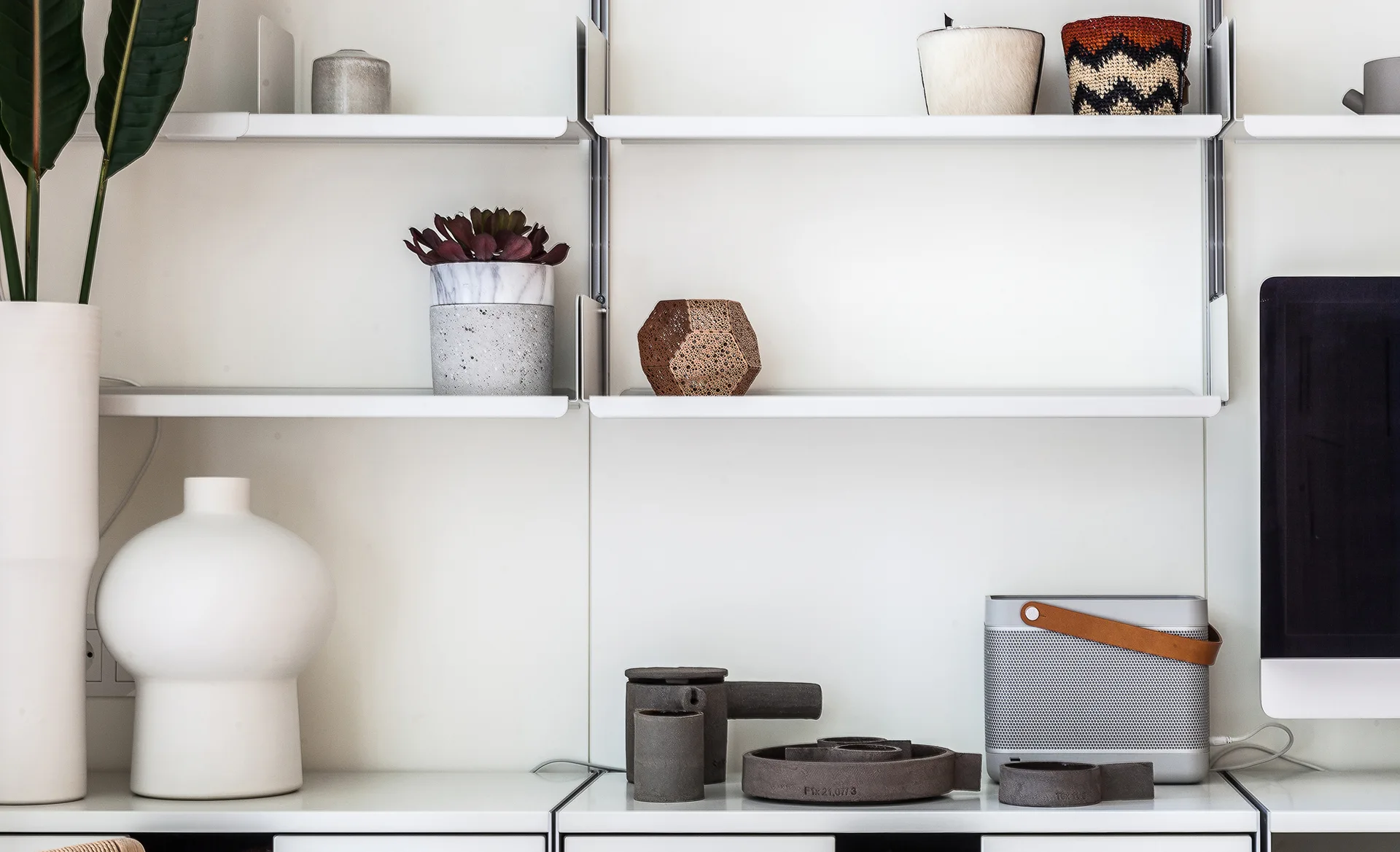 A minimalist white wall-mounted shelving unit is styled with geometric decor, ceramic vases, a succulent planter, and a portable speaker.