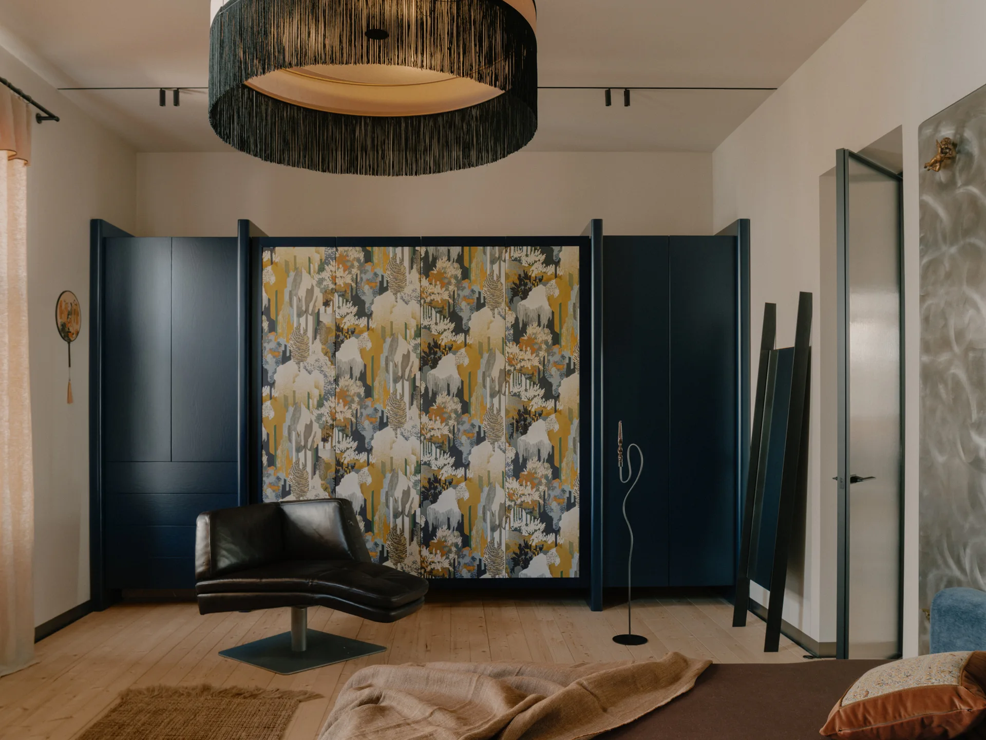 A bedroom featuring a large navy blue wardrobe with central patterned panels, a black leather swivel chair, and a large fringed pendant light overhead.