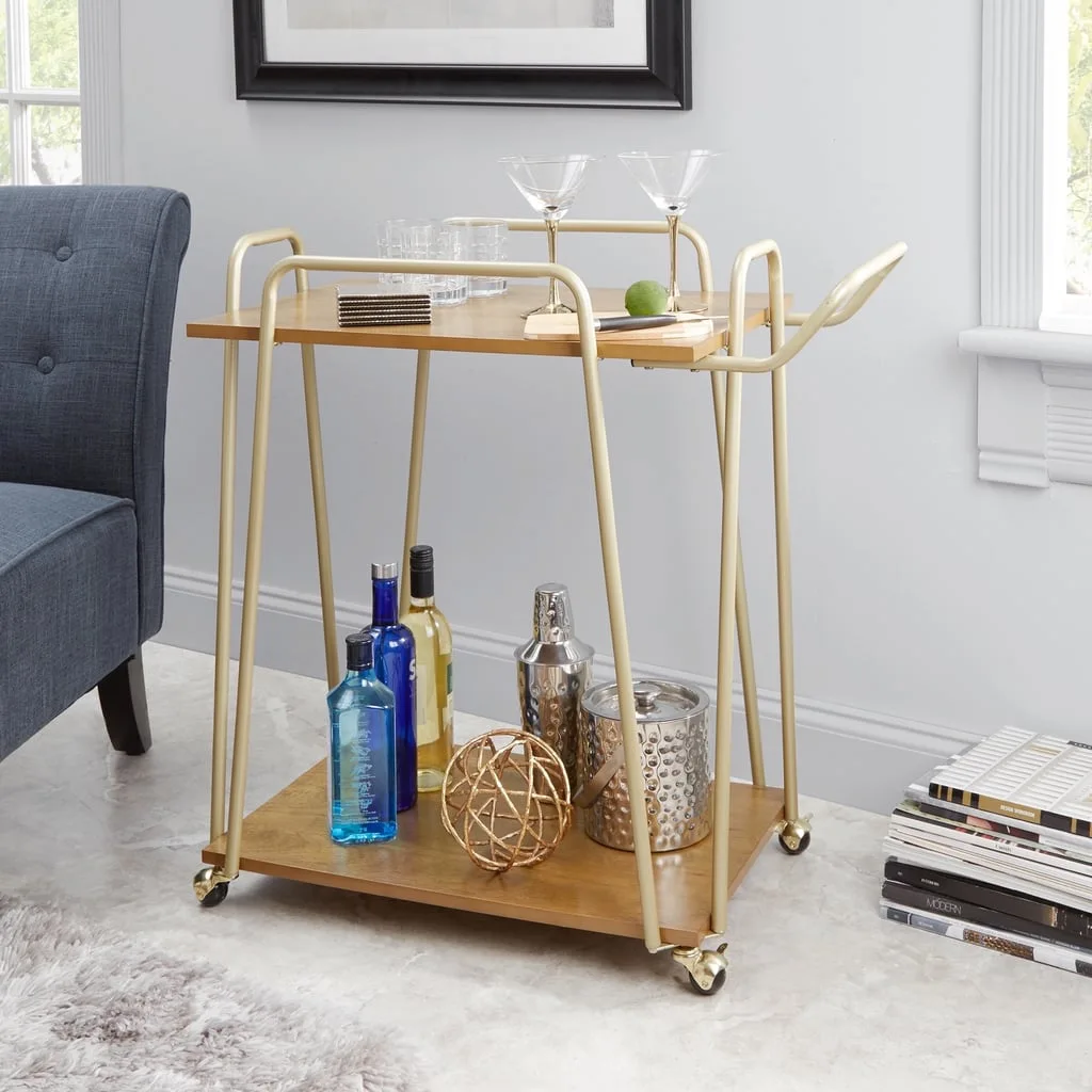 A two-tier gold and wood bar cart stocked with glassware and bottles sits on a marble floor next to a blue tufted armchair.
