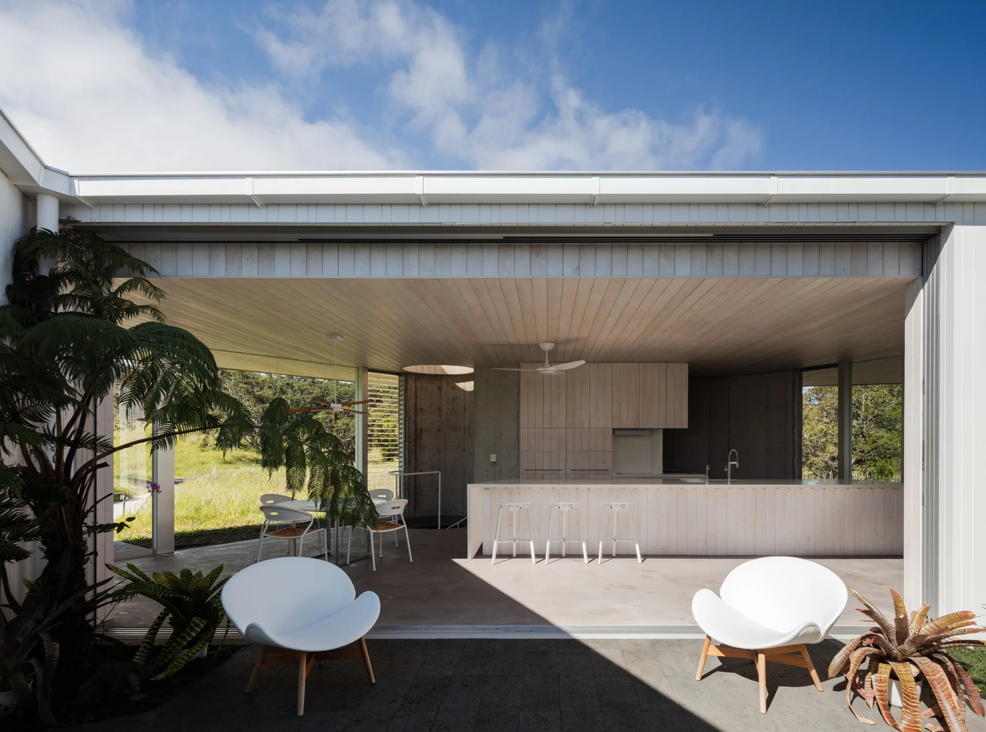 A modern covered patio with white armchairs features a seamless transition to an indoor kitchen with a white wood island and paneled ceiling.