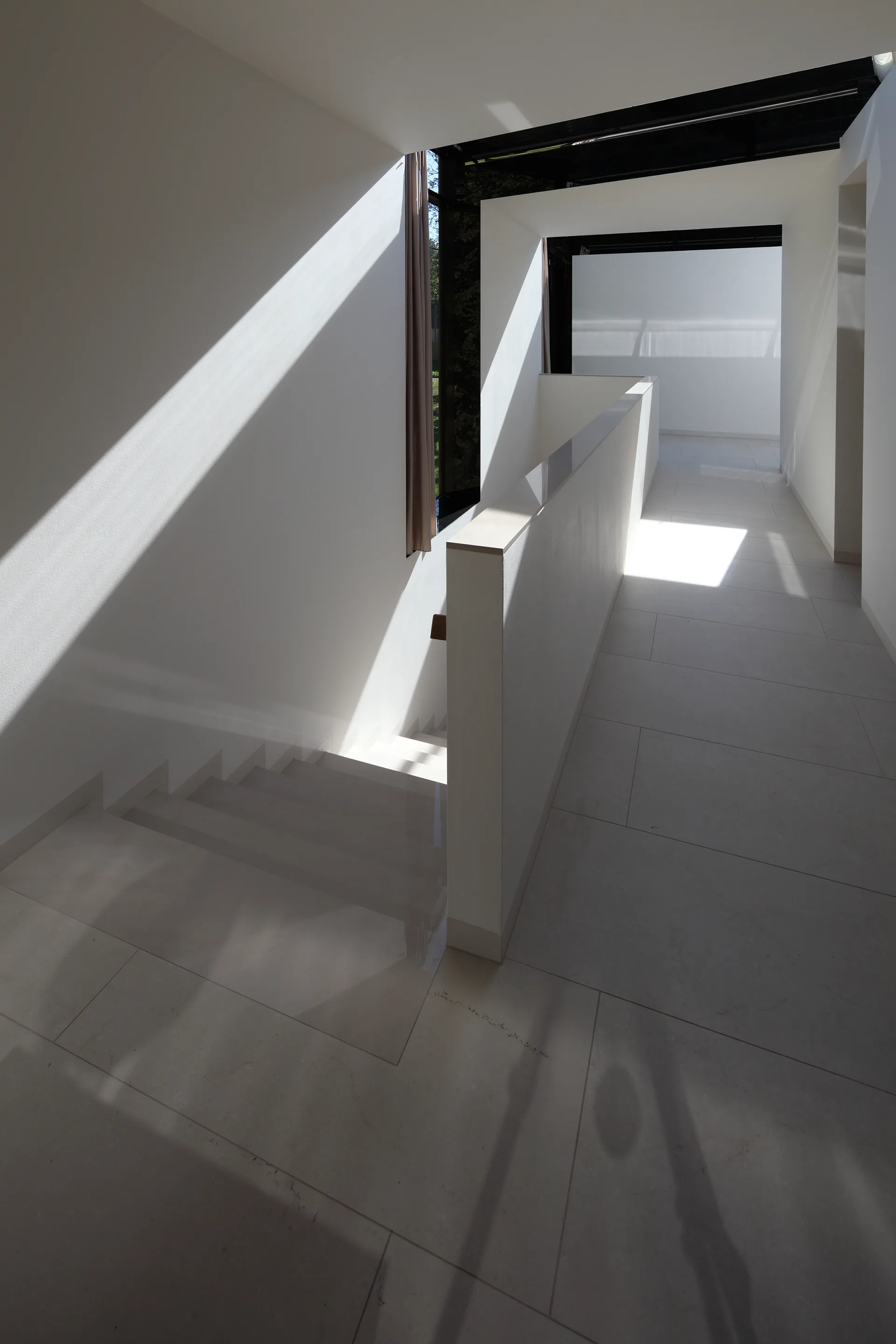 A minimalist white hallway and staircase featuring large-format grey tiles and strong geometric shadows from overhead natural light.