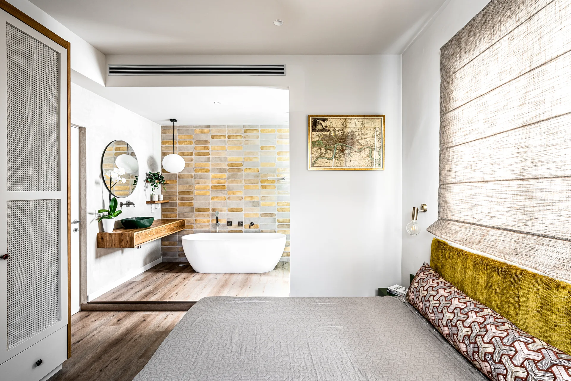 A modern bedroom looking into an open en-suite bathroom featuring a freestanding white tub, multi-toned tile wall, and a wood floating vanity.