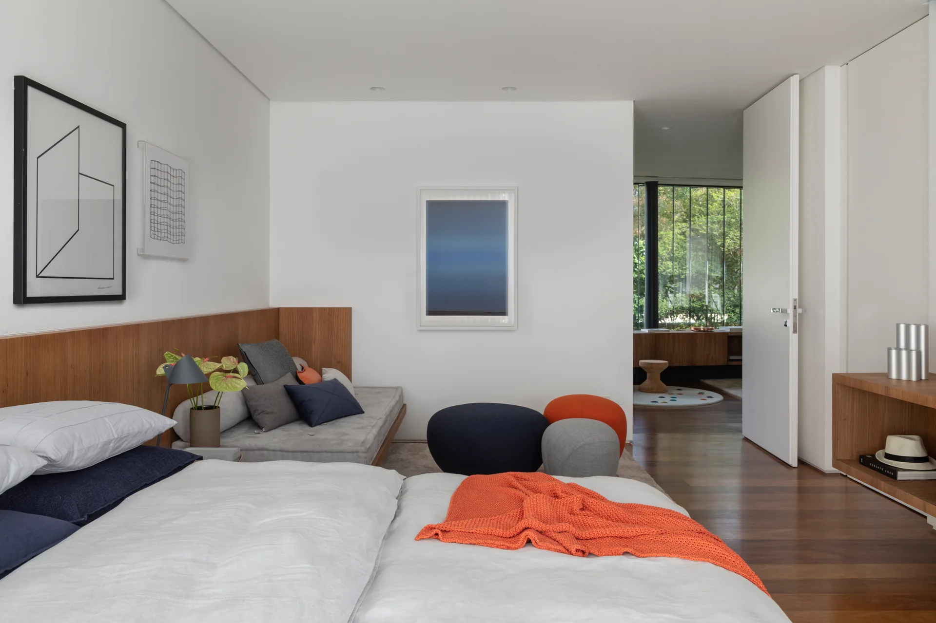 A modern bedroom features a white bed with an orange throw, a grey daybed against wood wall paneling, and colorful round ottomans.