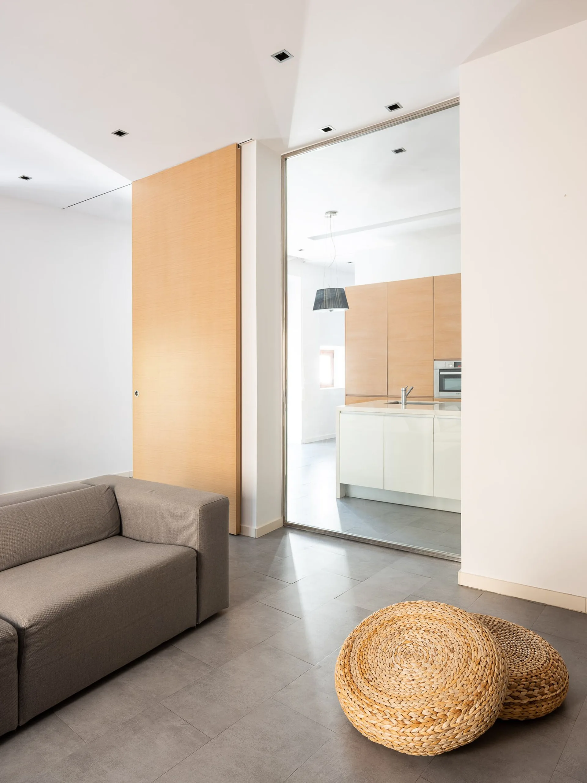 A minimalist living room with a gray sofa and woven floor cushions features a glass partition and wood sliding door next to a kitchen.