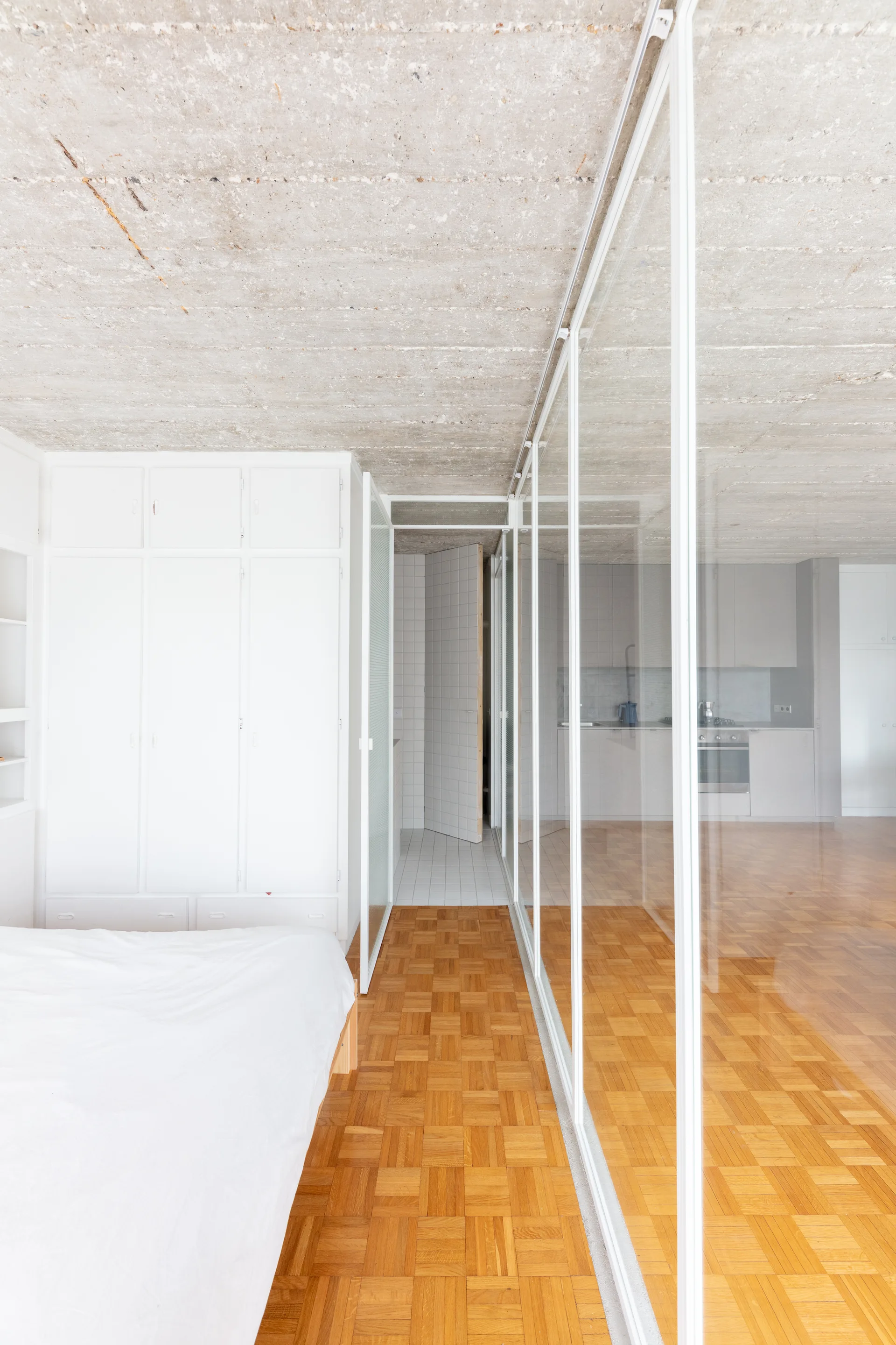 A minimalist bedroom features a white built-in wardrobe, a glass-paneled partition, warm wood parquet flooring, and an exposed concrete ceiling.