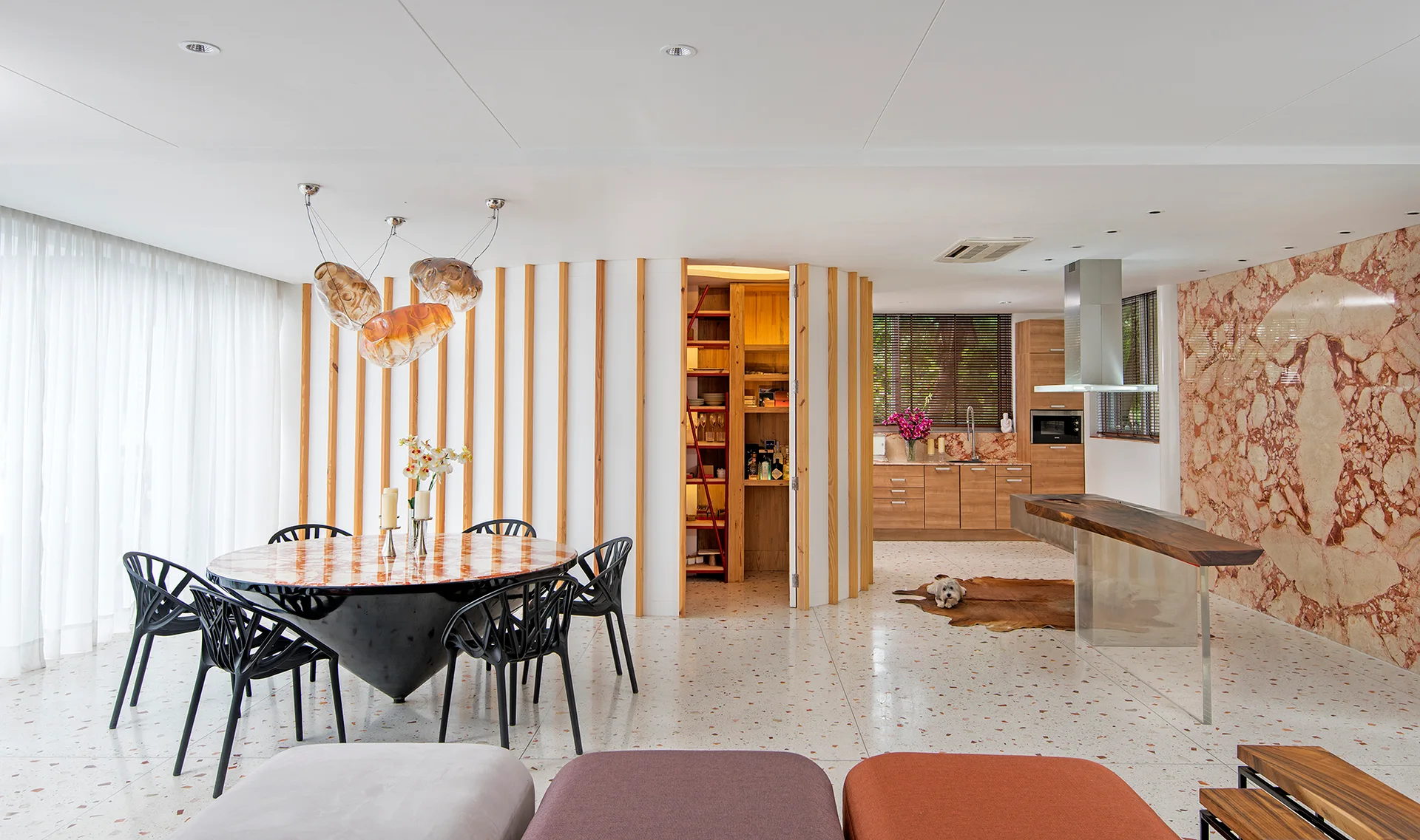 An open dining area features a round marble table, black geometric chairs, a curved vertical wood slat wall, and polished terrazzo flooring.